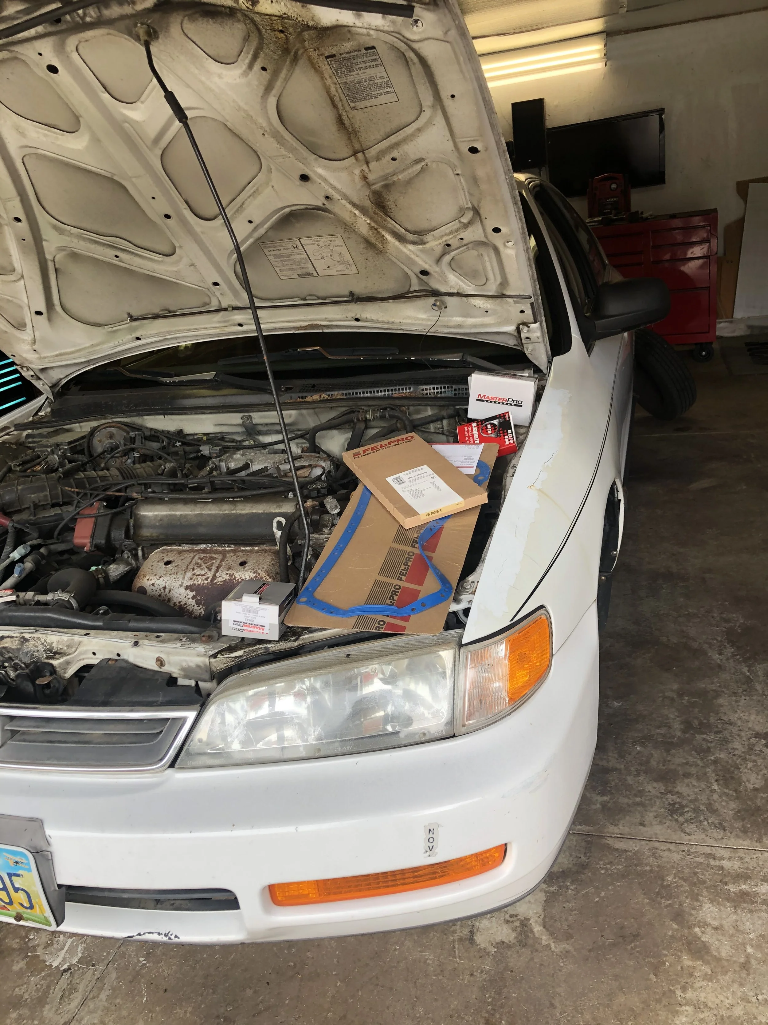 A white car with its hood open, showing the engine bay with various tools and parts placed on top of the engine.
