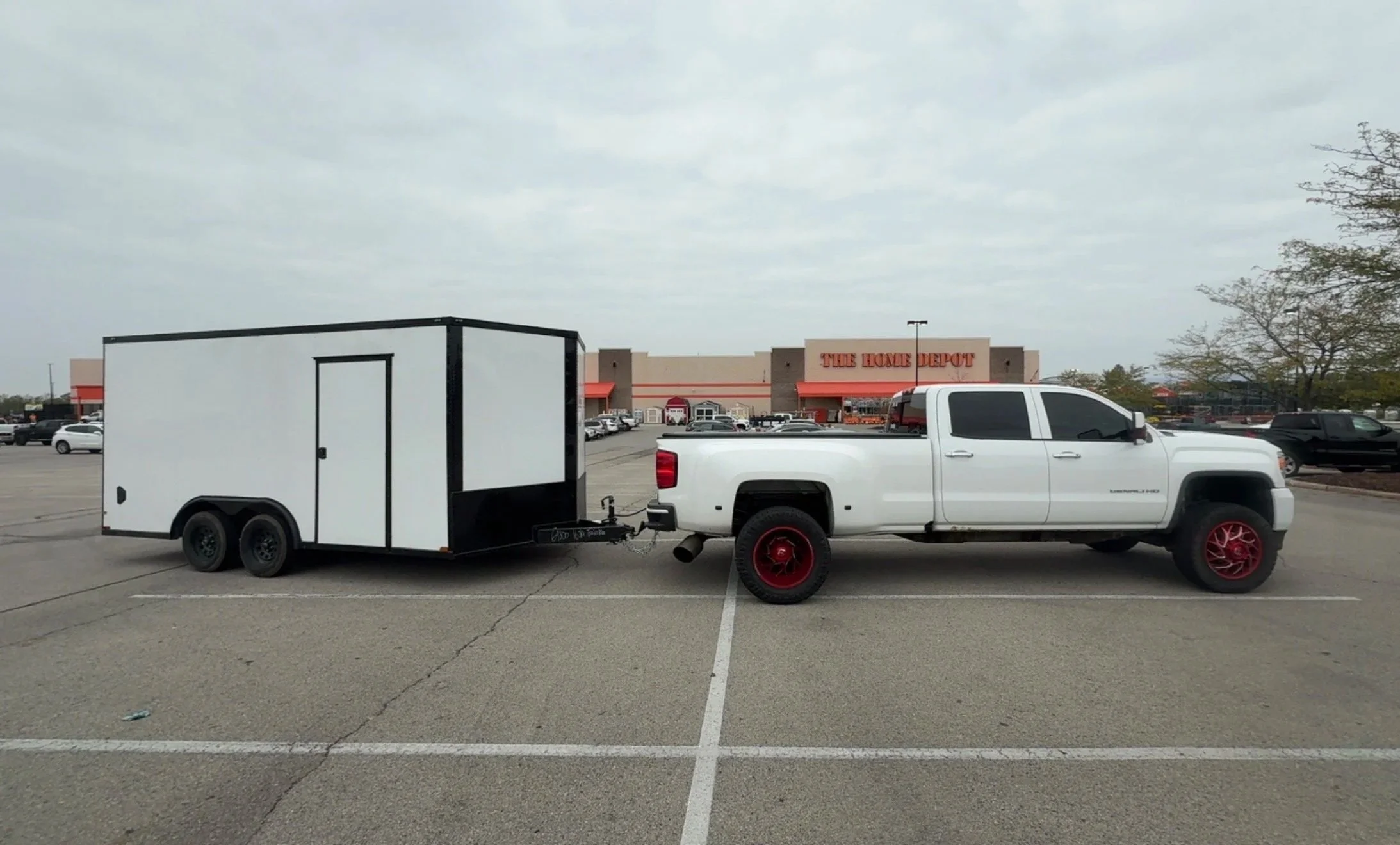 A white pickup truck with red wheels parked in a parking lot hitched to a large white enclosed trailer. In the background, there's a The Home Depot store and several parked cars on an overcast day.