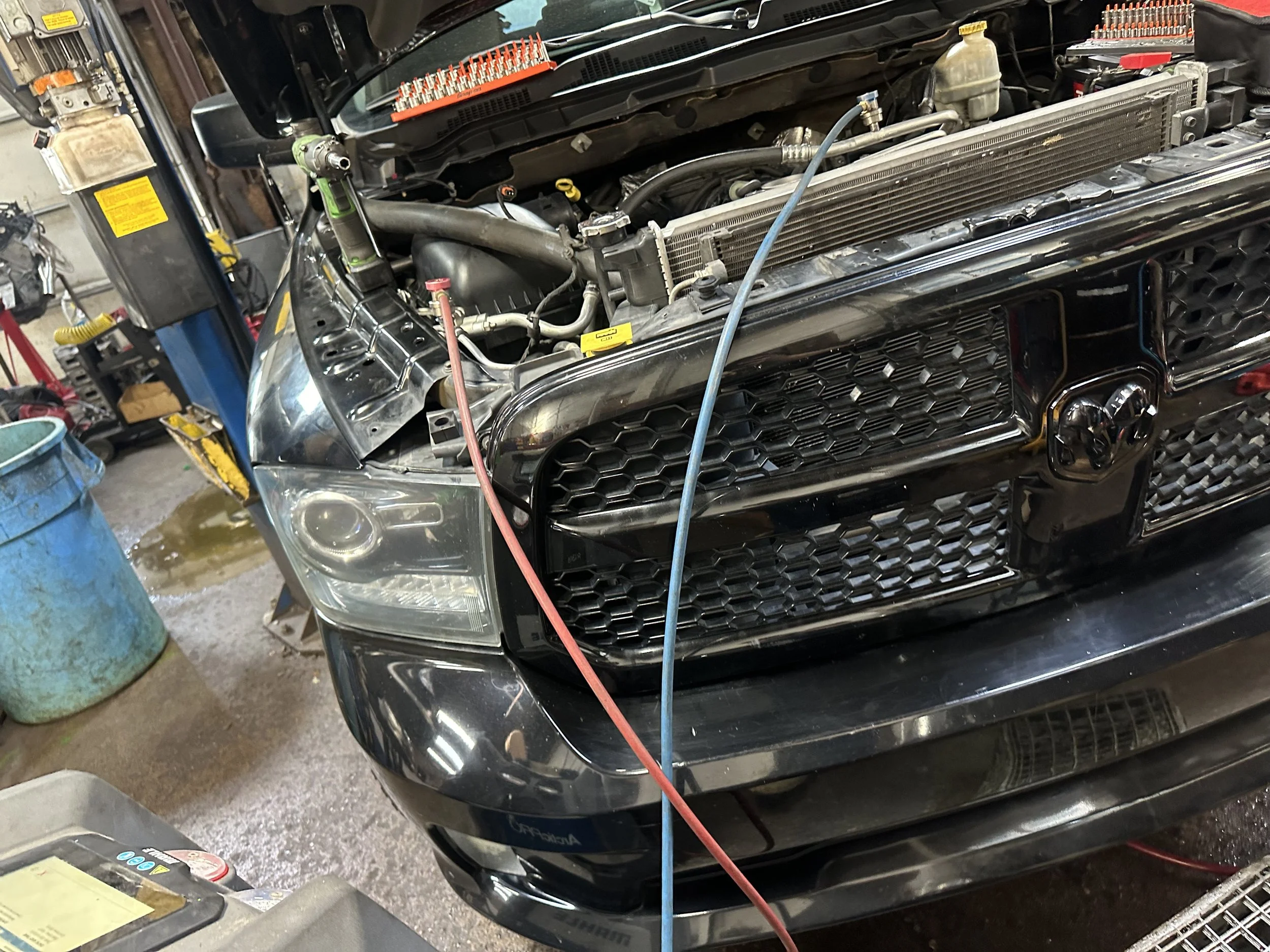Front view of a black pickup truck with its hood open, being serviced in a garage, with various tools and equipment visible around.