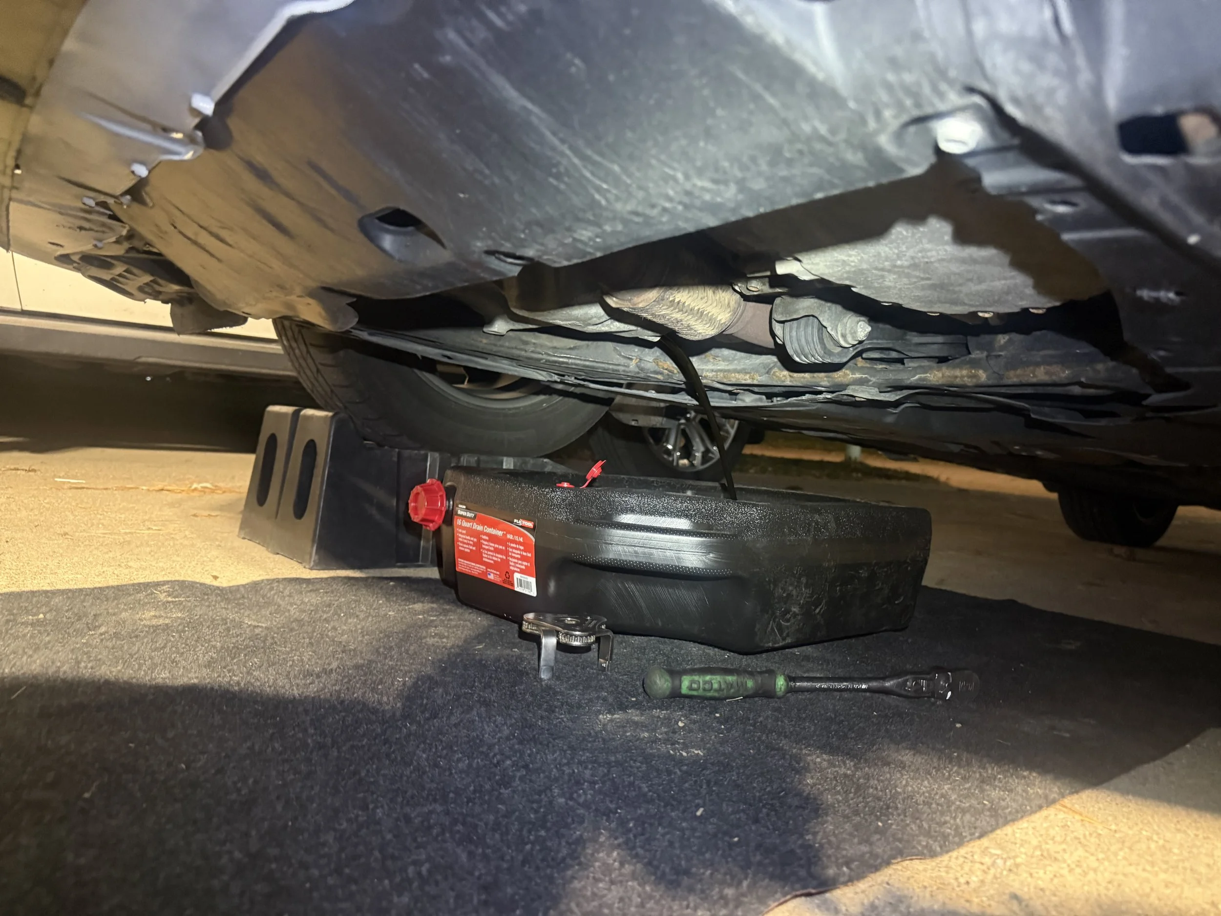 Underneath a car with the oil drain container and oil catch pan placed to catch draining oil. A wrench is on the ground beside the oil container, and blocks support the vehicle.