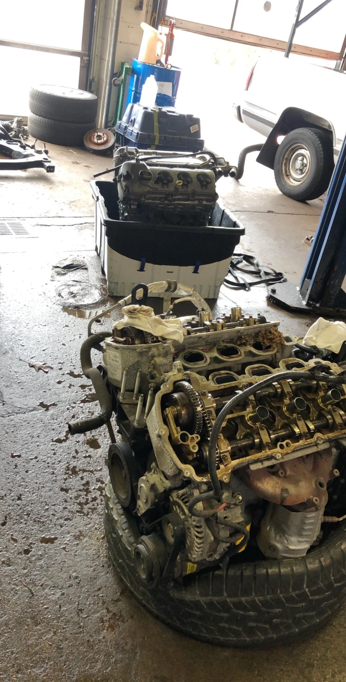 Automobile engine and cylinder head parts in a garage, with tools, spare tires, and a vehicle in background.