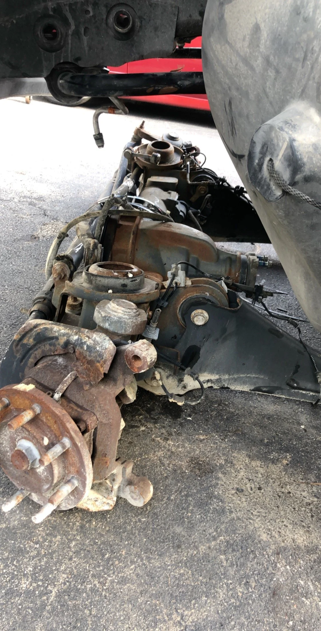 An old, rusty vehicle axle and differential assembly removed from a vehicle, lying on a rough asphalt surface.