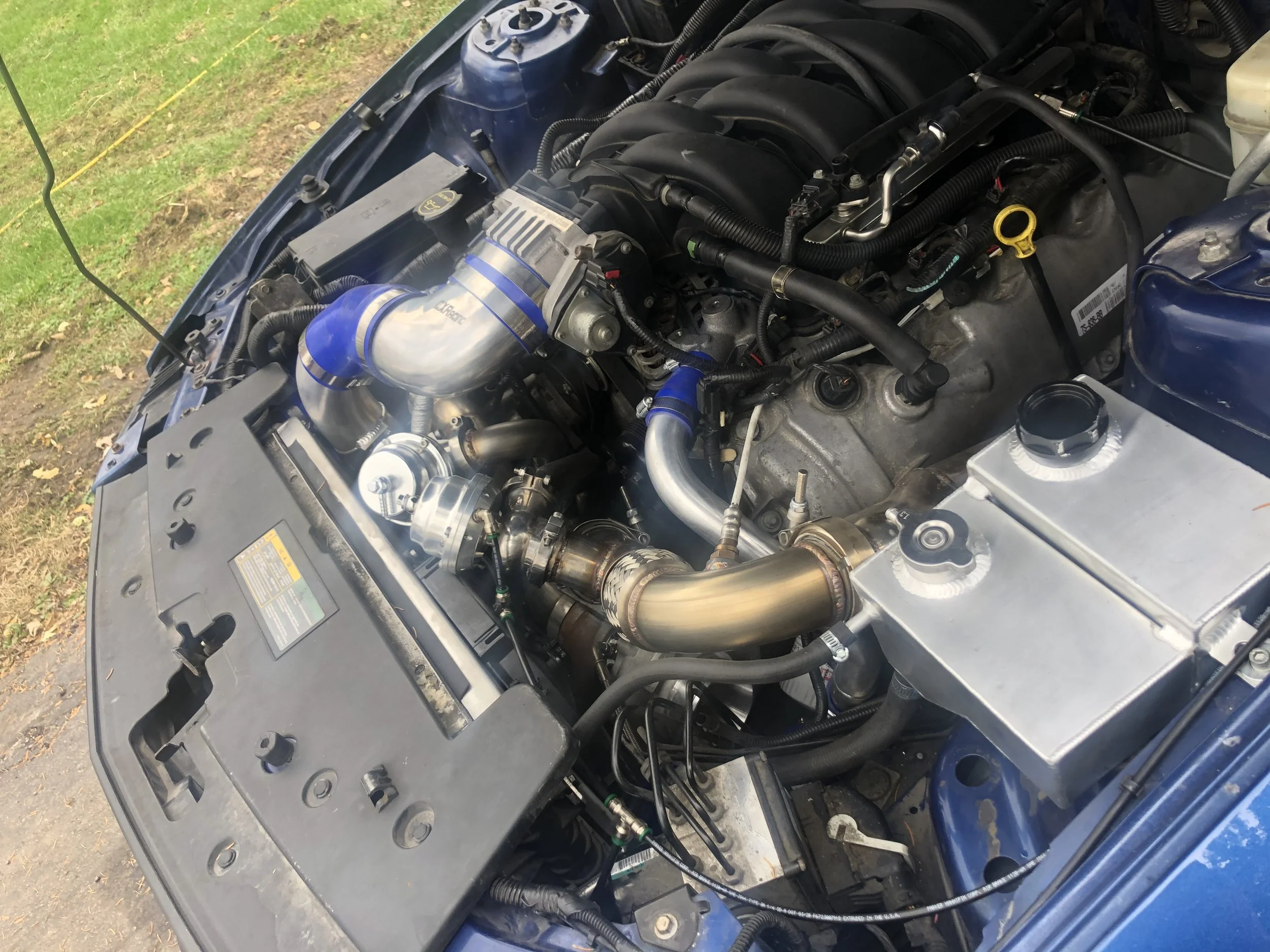Car engine bay with a large turbocharger installed, featuring blue silicone hoses and a golden-colored piping, along with various metal components, electrical wiring, and fluid reservoirs.