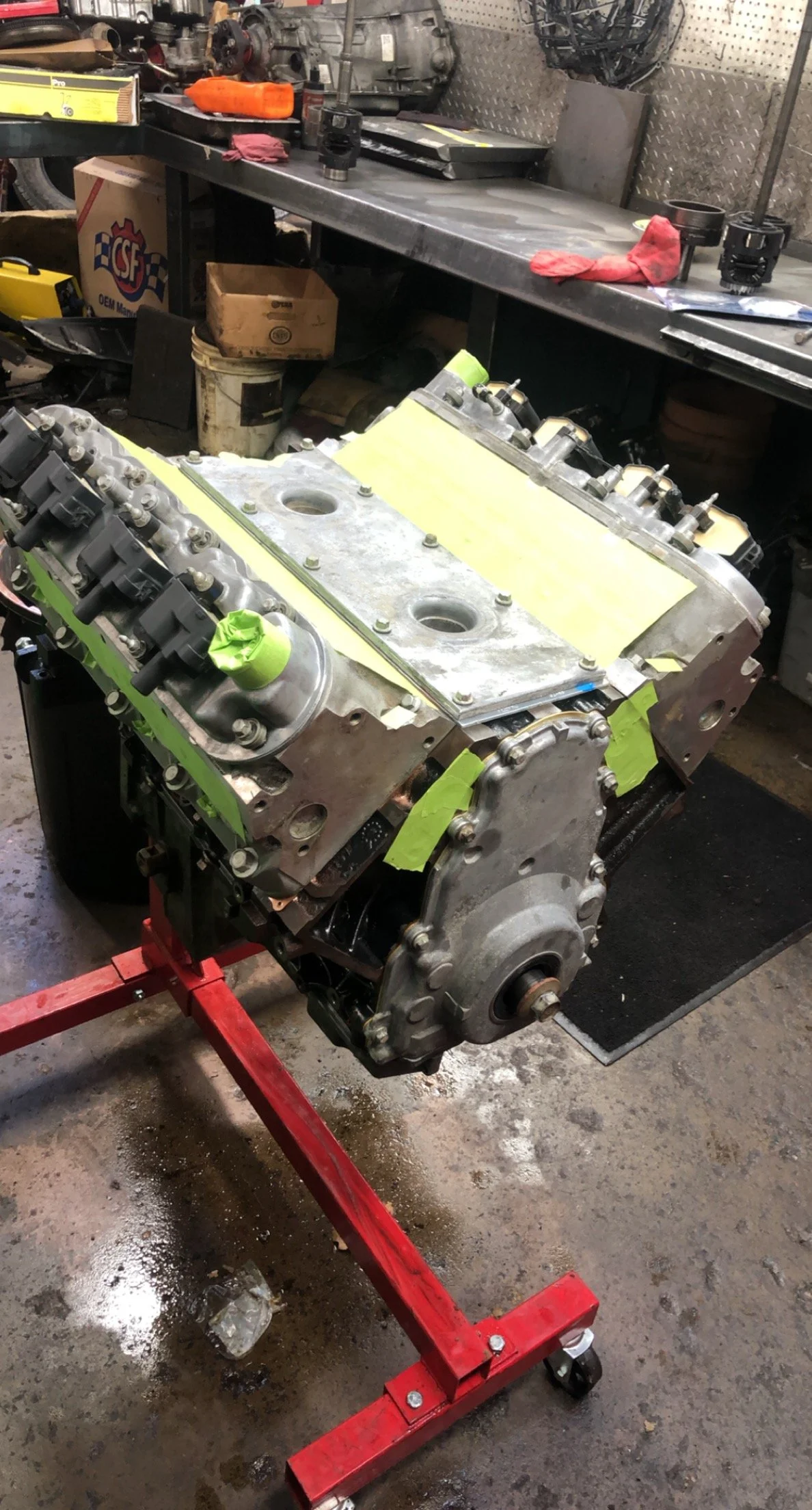 An engine block mounted on a red engine stand in a workshop with tools and parts on a workbench in the background.