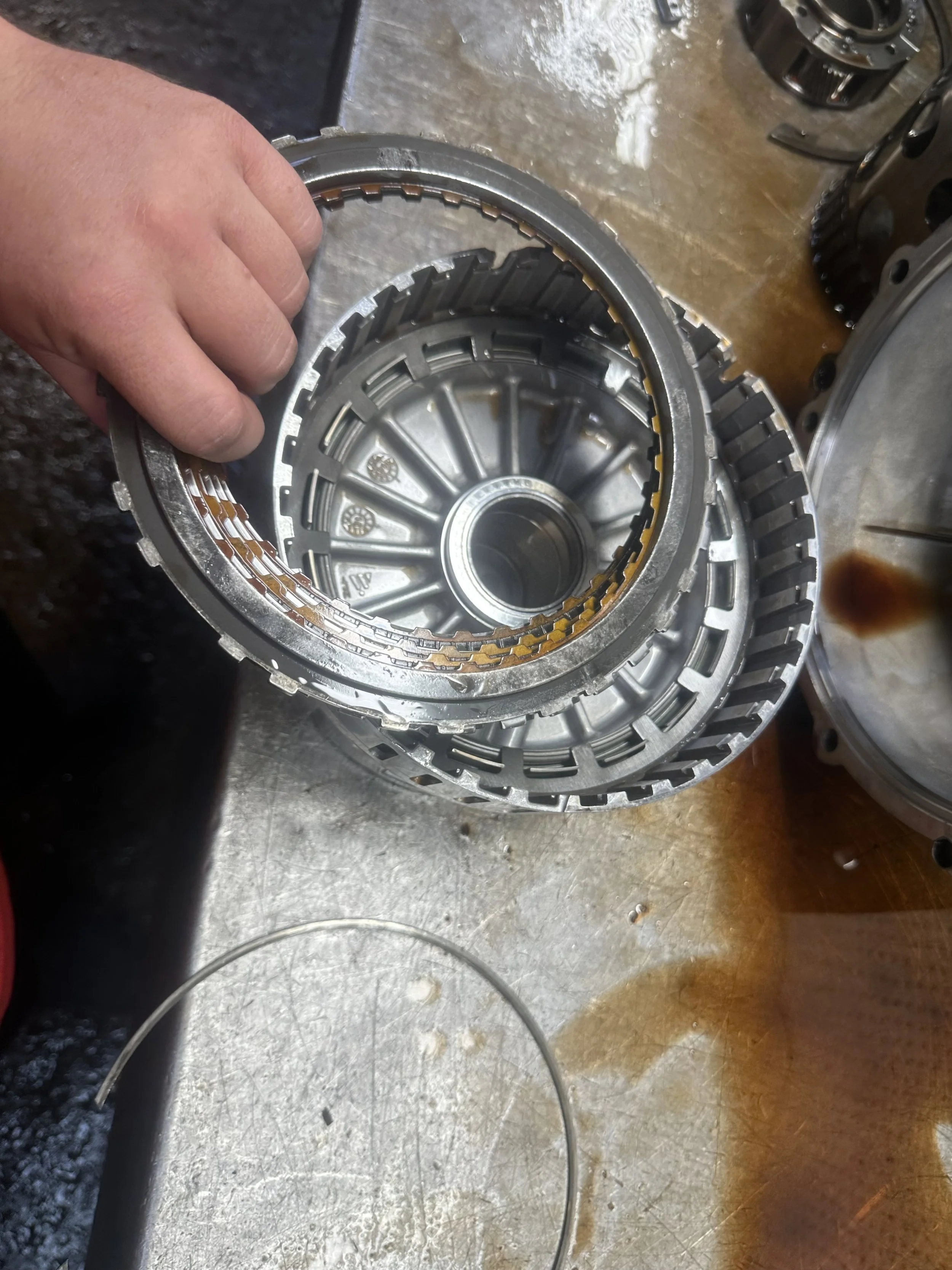 Close-up of a person holding a metallic automotive transmission component, with tools and other car parts visible on a workbench.