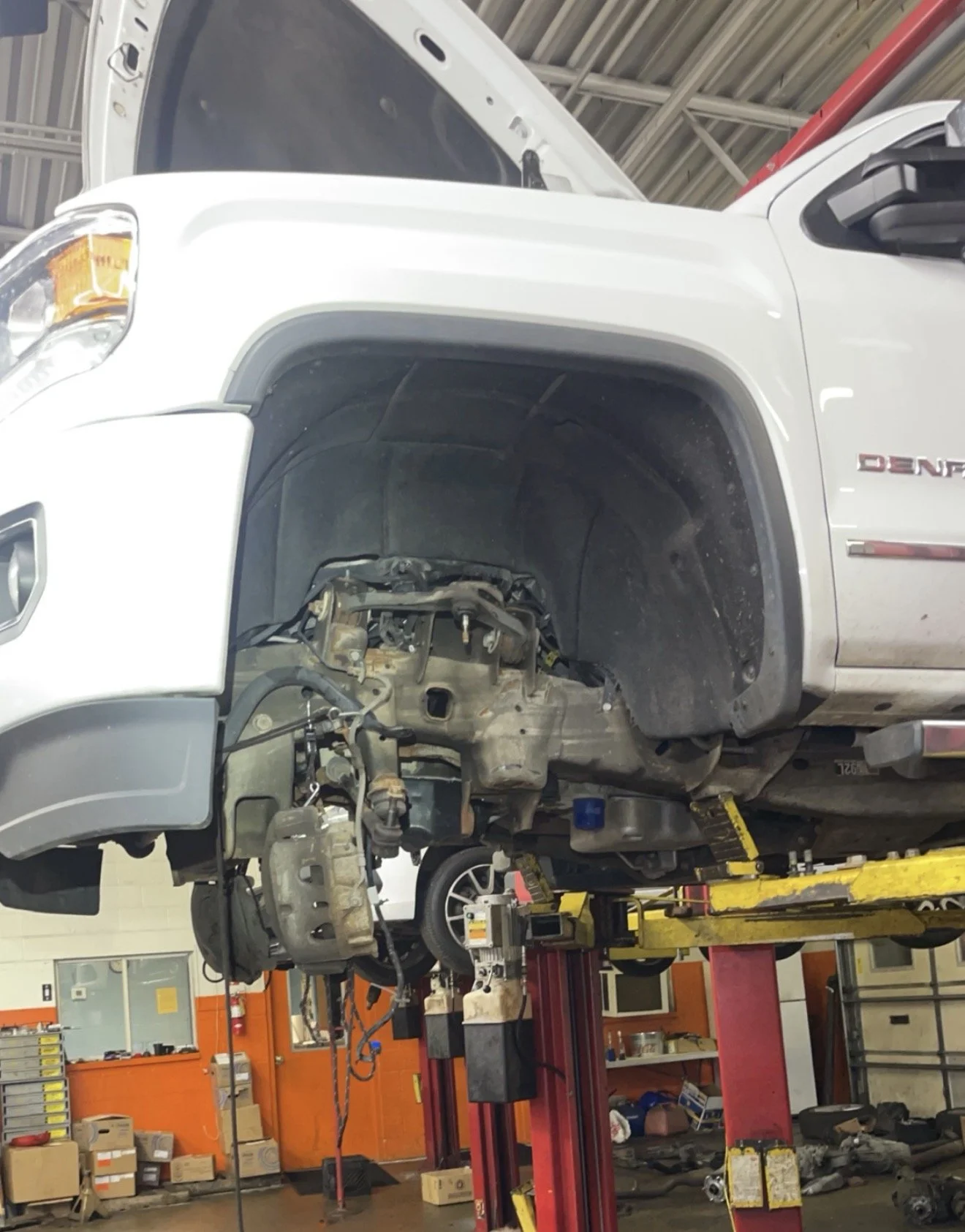 The front of a white GMC Denali truck is lifted on a red hydraulic car lift inside an auto repair shop, with the wheel removed and the suspension and brake components visible.