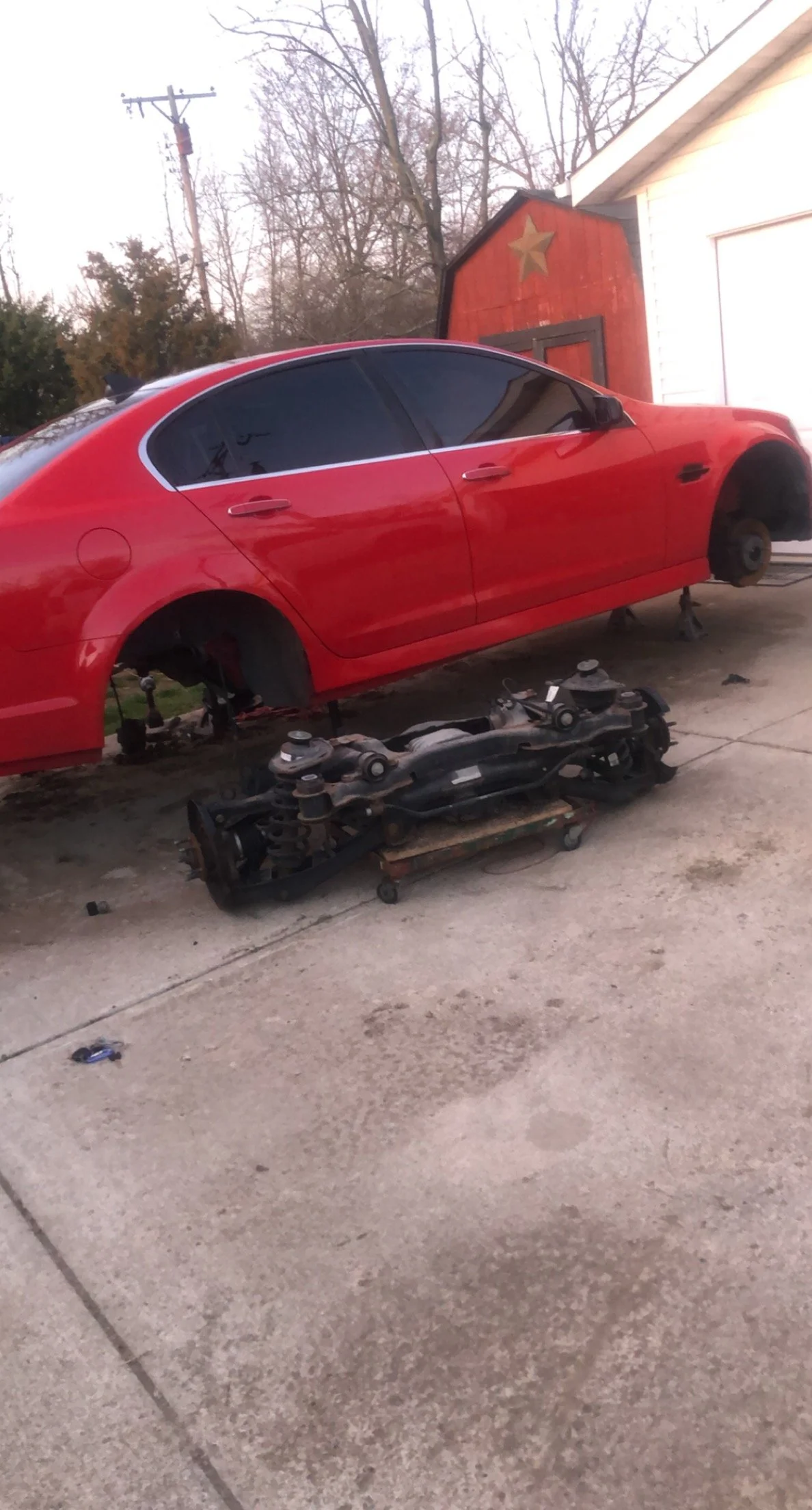 A red sedan car missing its wheels is parked on the driveway, with the wheels and suspension assembly placed on a dolly underneath the car.