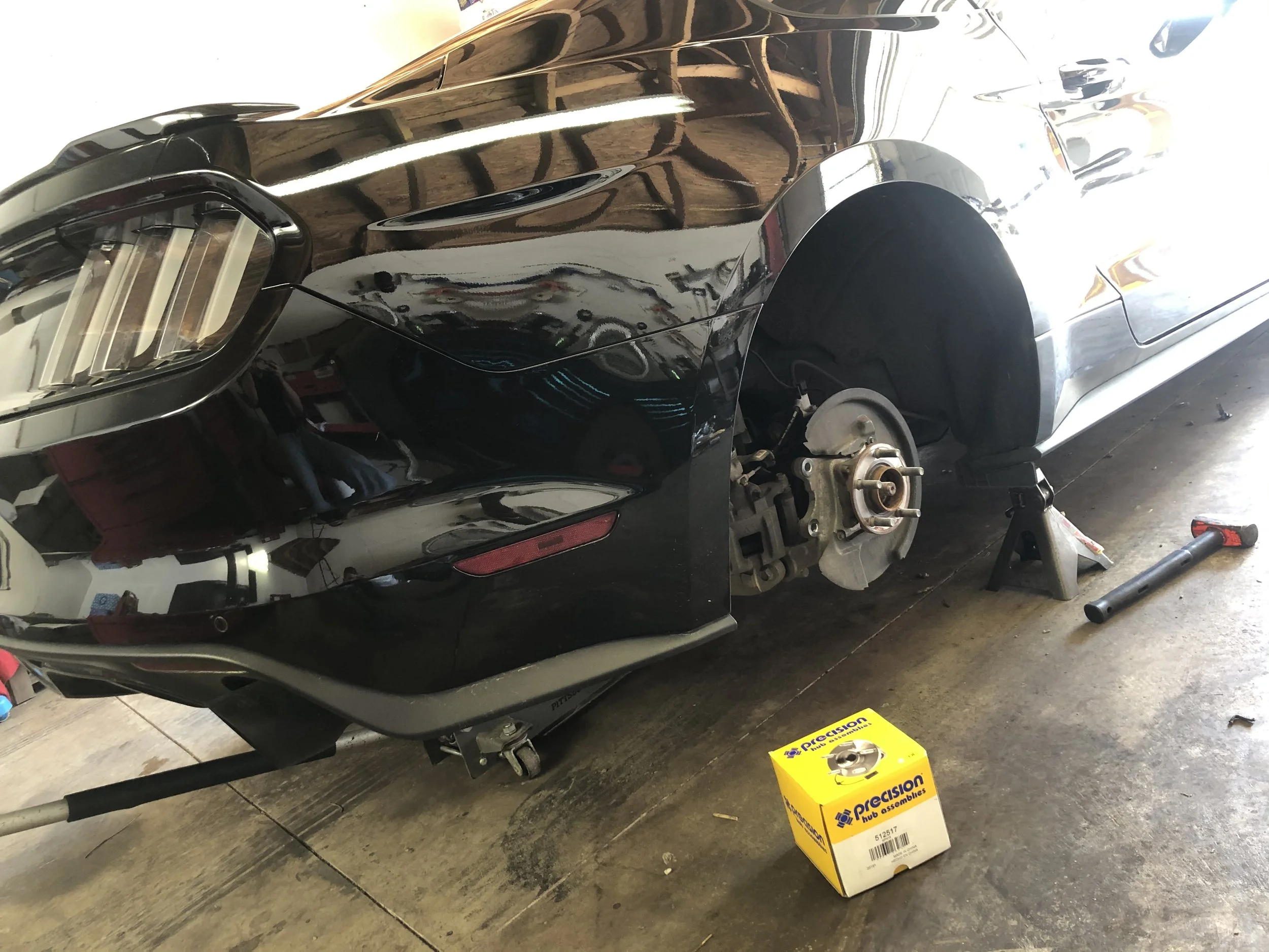 A black car is elevated on jack stands with its front wheel removed, exposing the brake assembly. The car is in a garage, and various tools and car parts, including a yellow box labeled 'Precision' and a red-handled tool, are visible on the concrete floor.