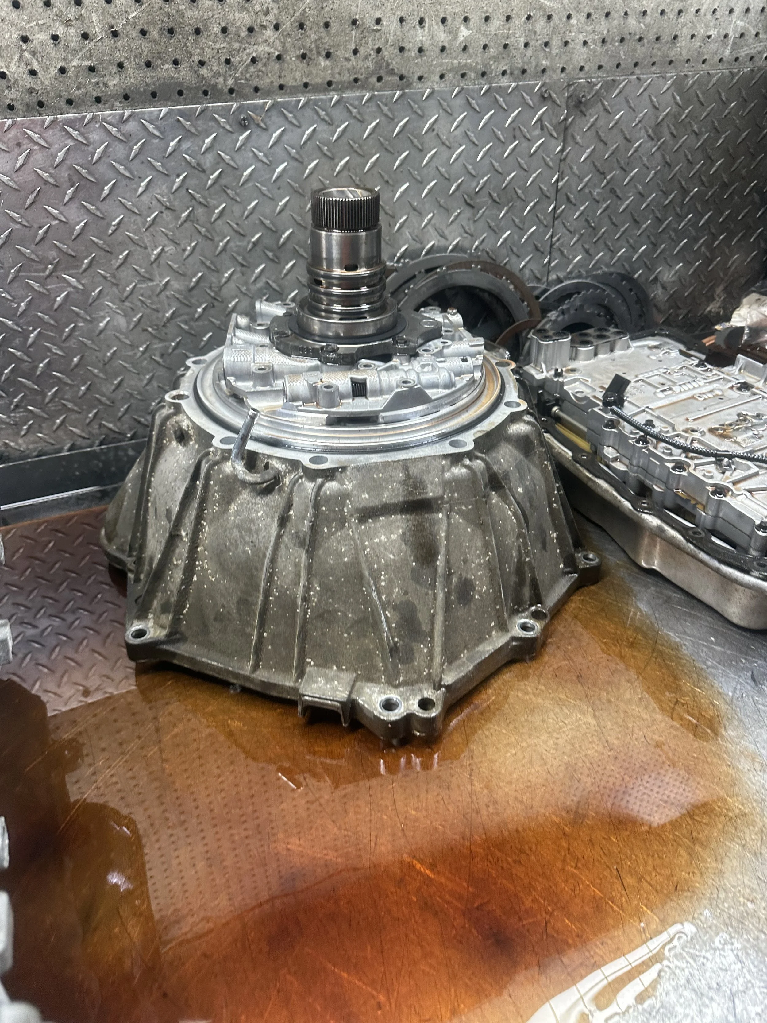 Automotive transmission parts on a workbench, including a dirty transmission casing, a metal piece with a shaft, and a metallic component, with tools and parts in the background.