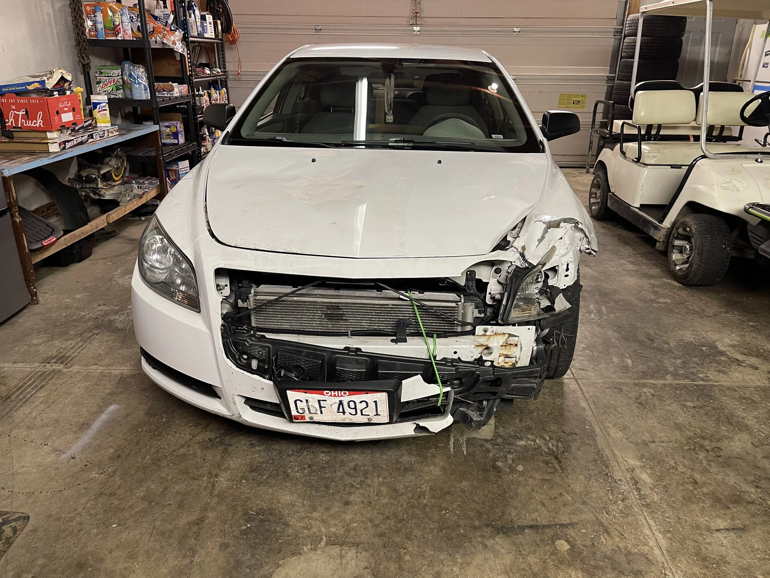White car with front-end damage in a garage, missing front bumper and grille, with a Ohio license plate reading 'GLF 4921'.