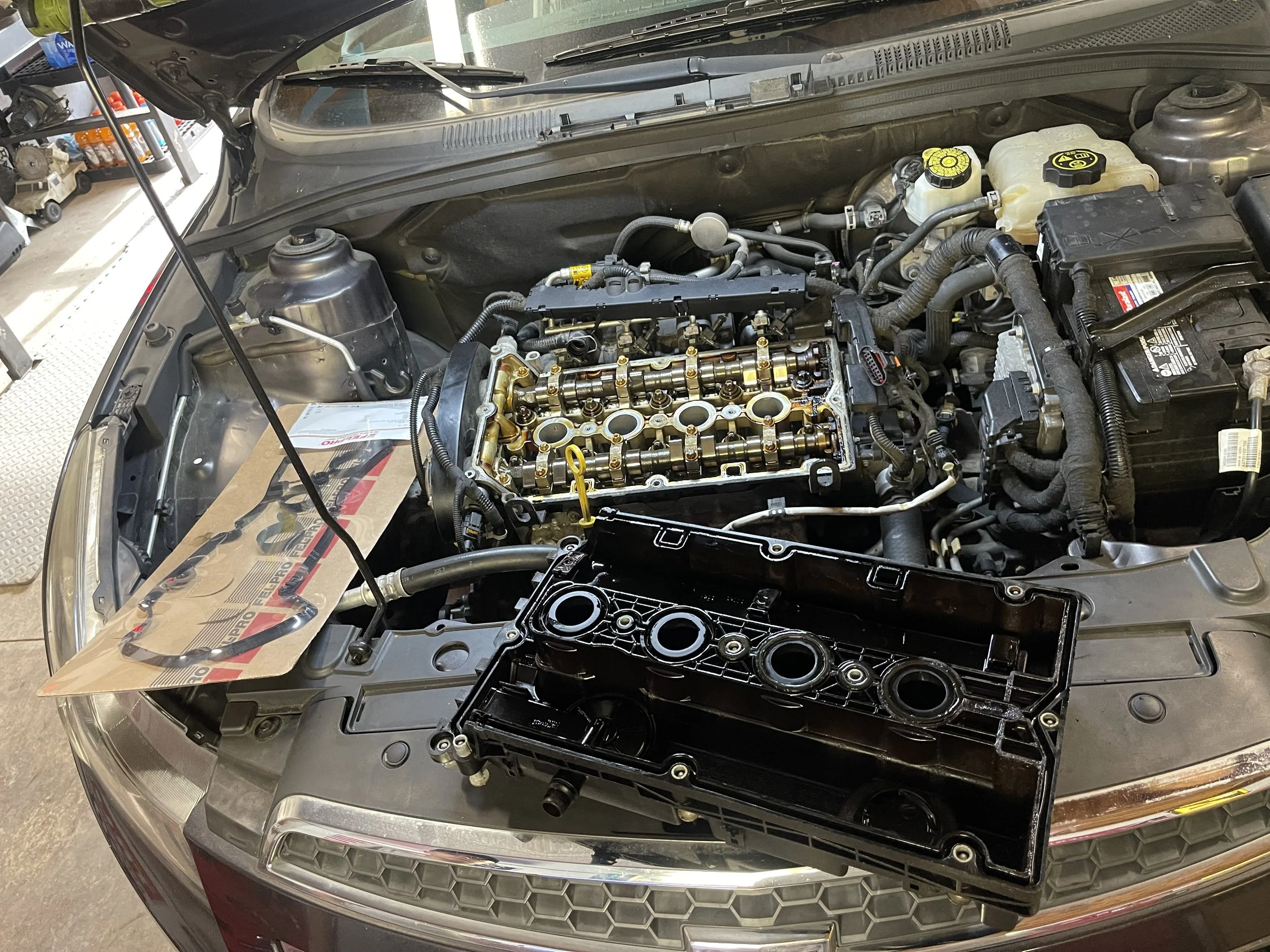 Open car hood revealing engine with valve cover removed, exposing camshaft and valves, with engine components and tools nearby in a garage.