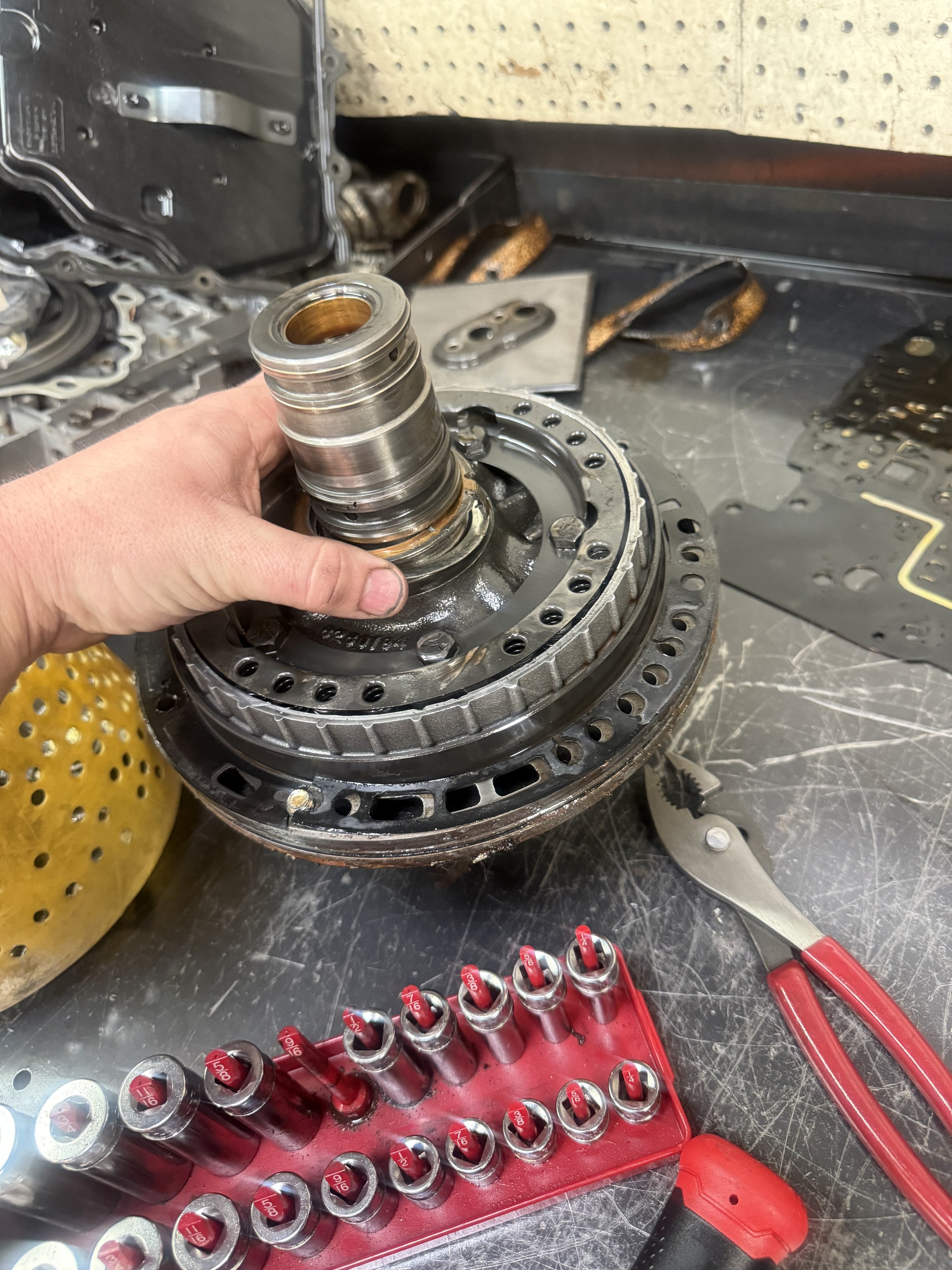 Close-up of a mechanic's workspace with a person holding a disassembled component of a machine or engine over a workbench scattered with tools, parts, and machinery.