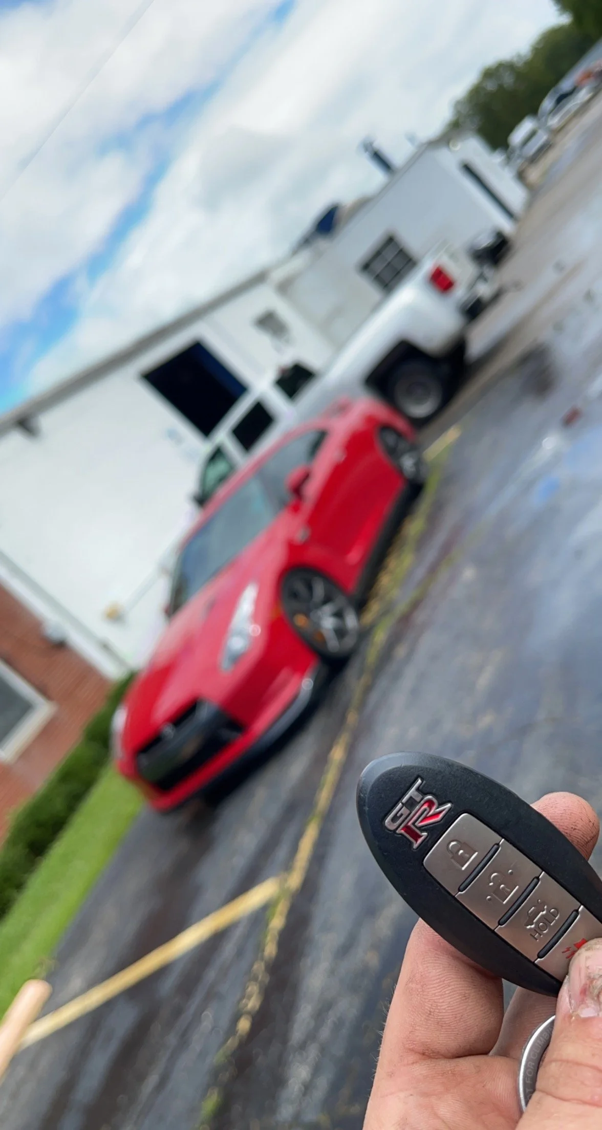 A person holding a car key fob with a red sports car, a white truck, and a house in the background. The scene is outdoors on a cloudy day.