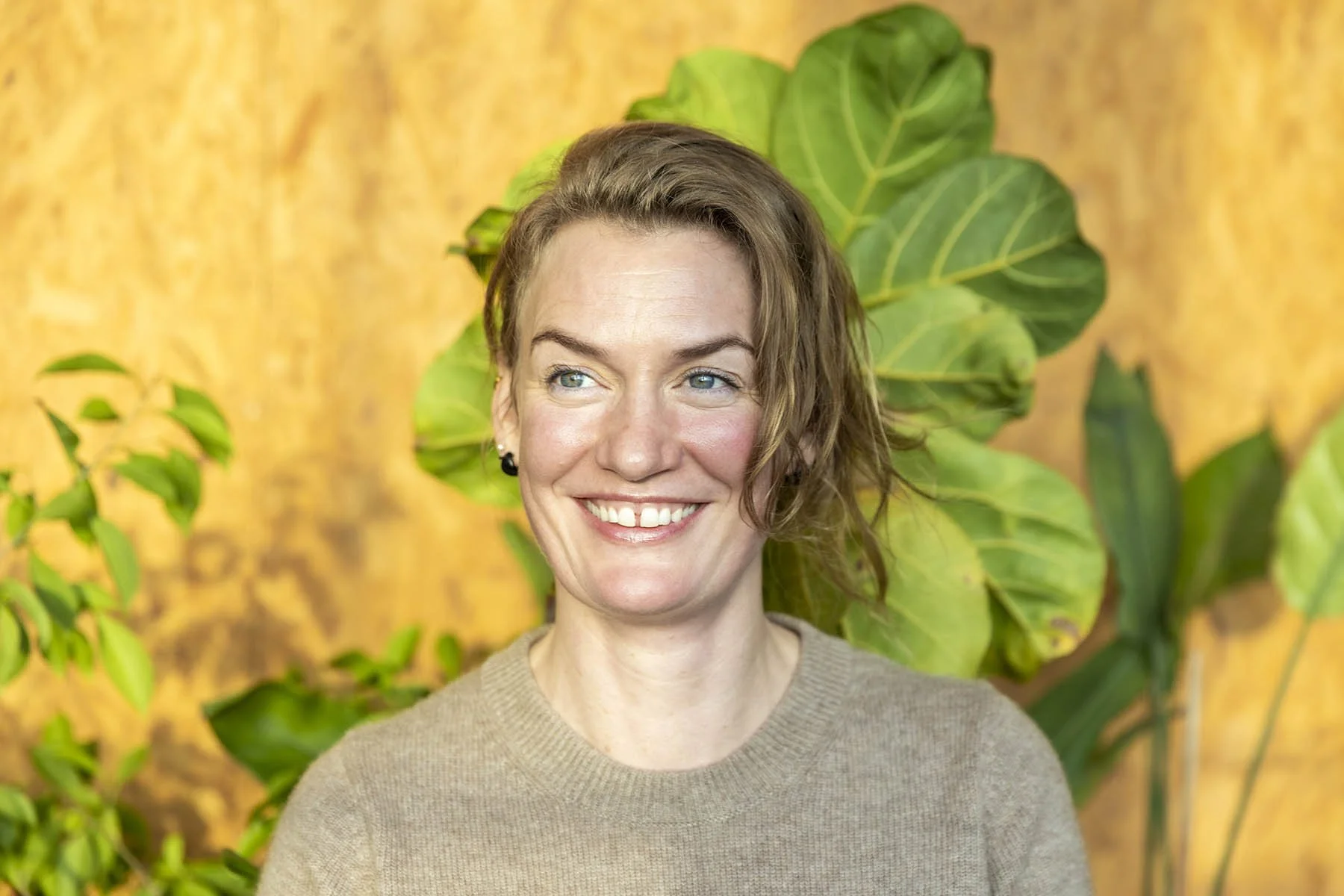 Et smilende kvinden med kort brunt hår, iført en beige sweater, står foran en grøn plante og en lysegul baggrund.