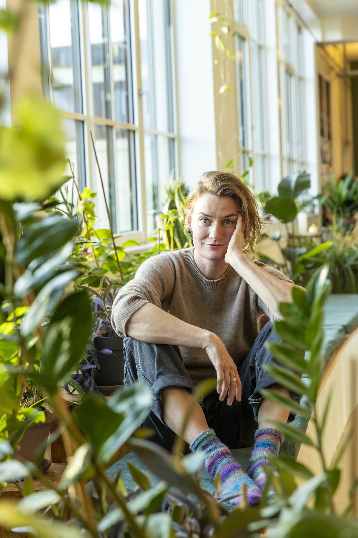 Kvinde sidder blandt grønne planter i et rum med store vinduer, iført en grisefarvet sweater, jeans og farverige sokker.