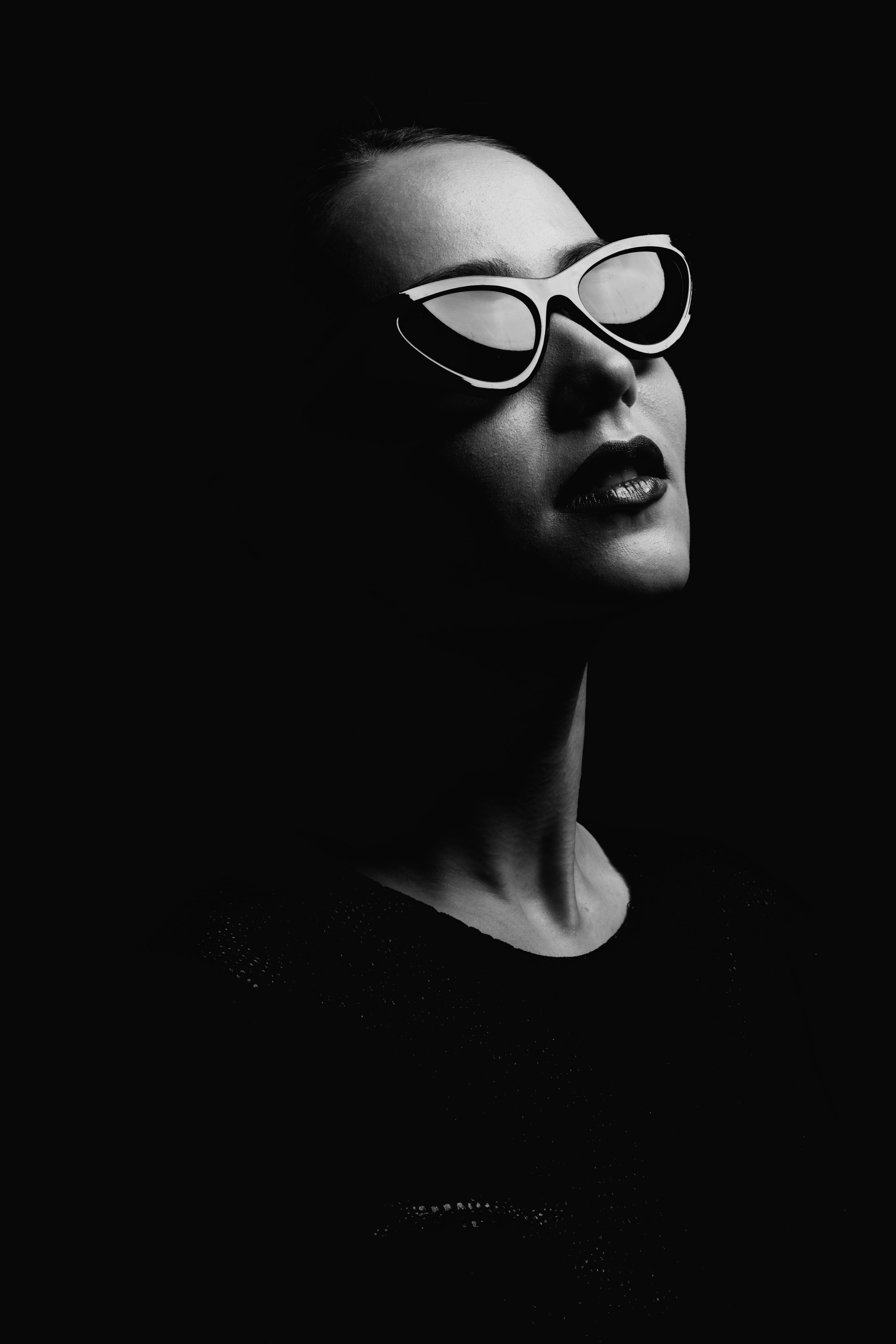 Black and white portrait of a woman wearing oversized sunglasses and dark lipstick, with high contrast lighting emphasizing her facial features.