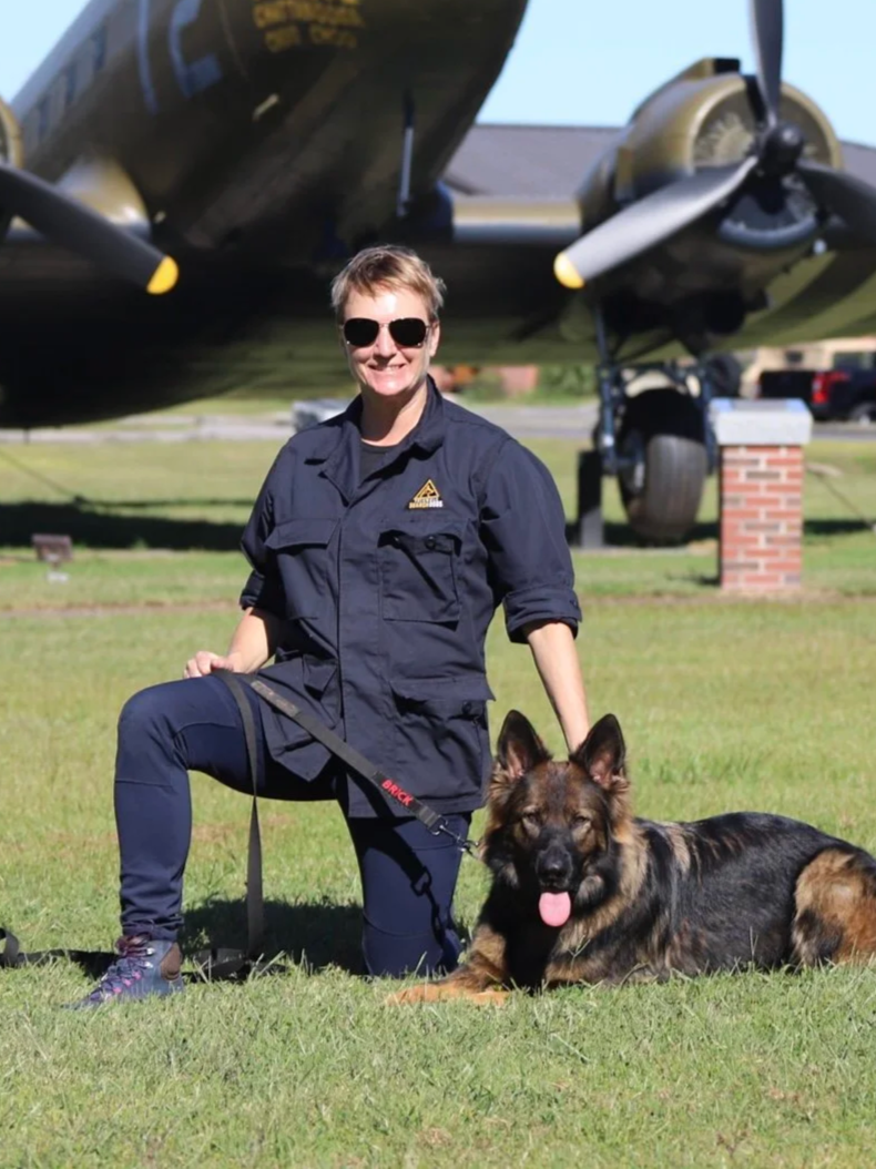 A woman with short hair and sunglasses kneeling on grass, smiling, with a German Shepherd dog lying beside her. They are outdoors in front of a vintage military aircraft.