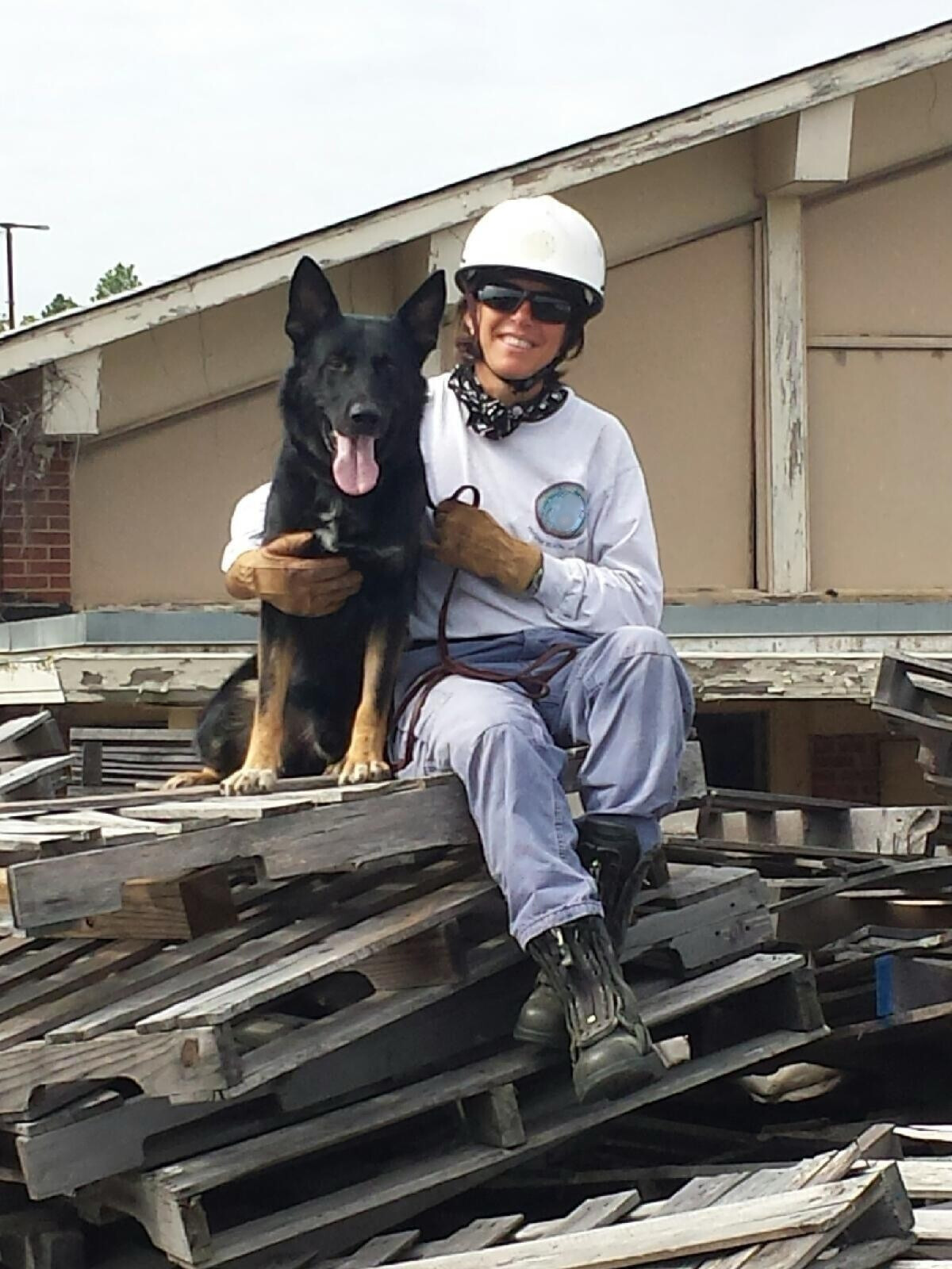 A woman in construction gear, sitting on a pile of wood with a black and tan dog, both looking at the camera, outside a building with a weathered roof.