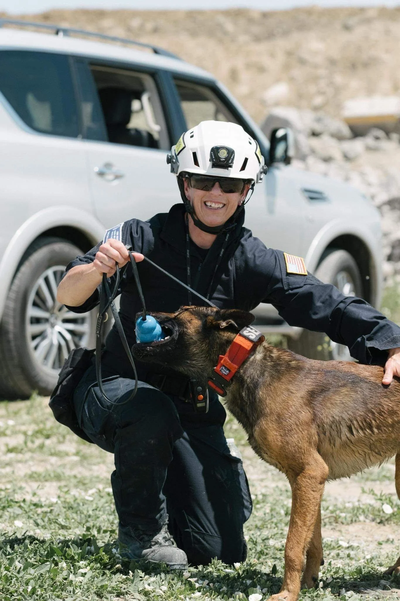A rescue worker kneeling on the ground with a police dog, both smiling. The rescue worker is wearing a helmet and sunglasses, and is holding a leash attached to the dog, which is licking a blue toy in the worker's hand. There is a gray vehicle in the background.
