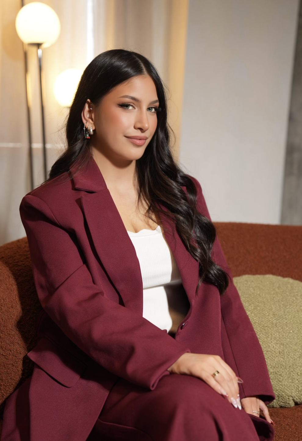 A woman with long dark hair and light makeup sitting on a brown sofa, wearing a maroon blazer and pants with a white top, in a room with beige curtains and a floor lamp with warm lights.
