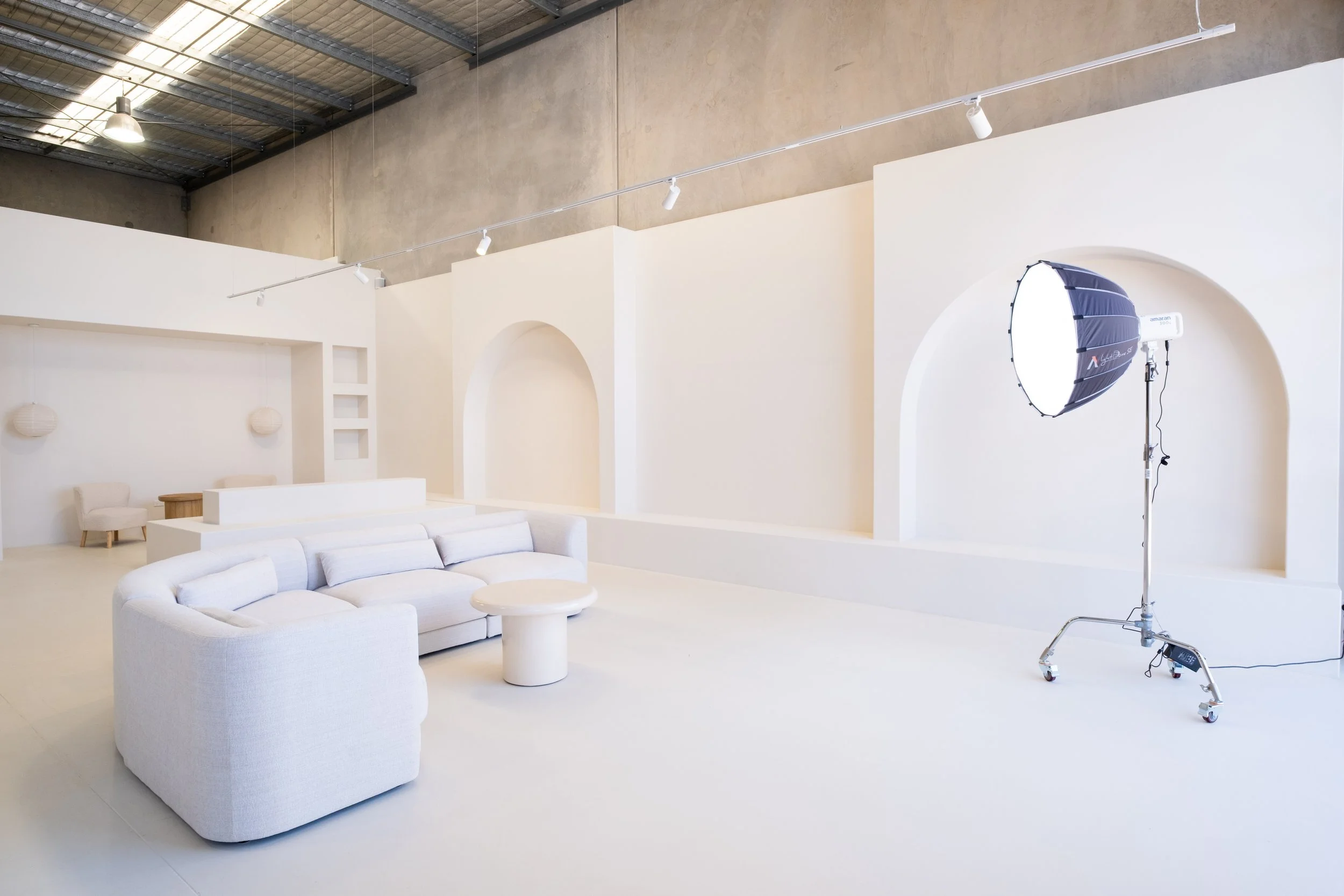 A large photo studio with white walls and floor, studio lighting equipment, a white sectional sofa, a round coffee table, and minimalistic decor.