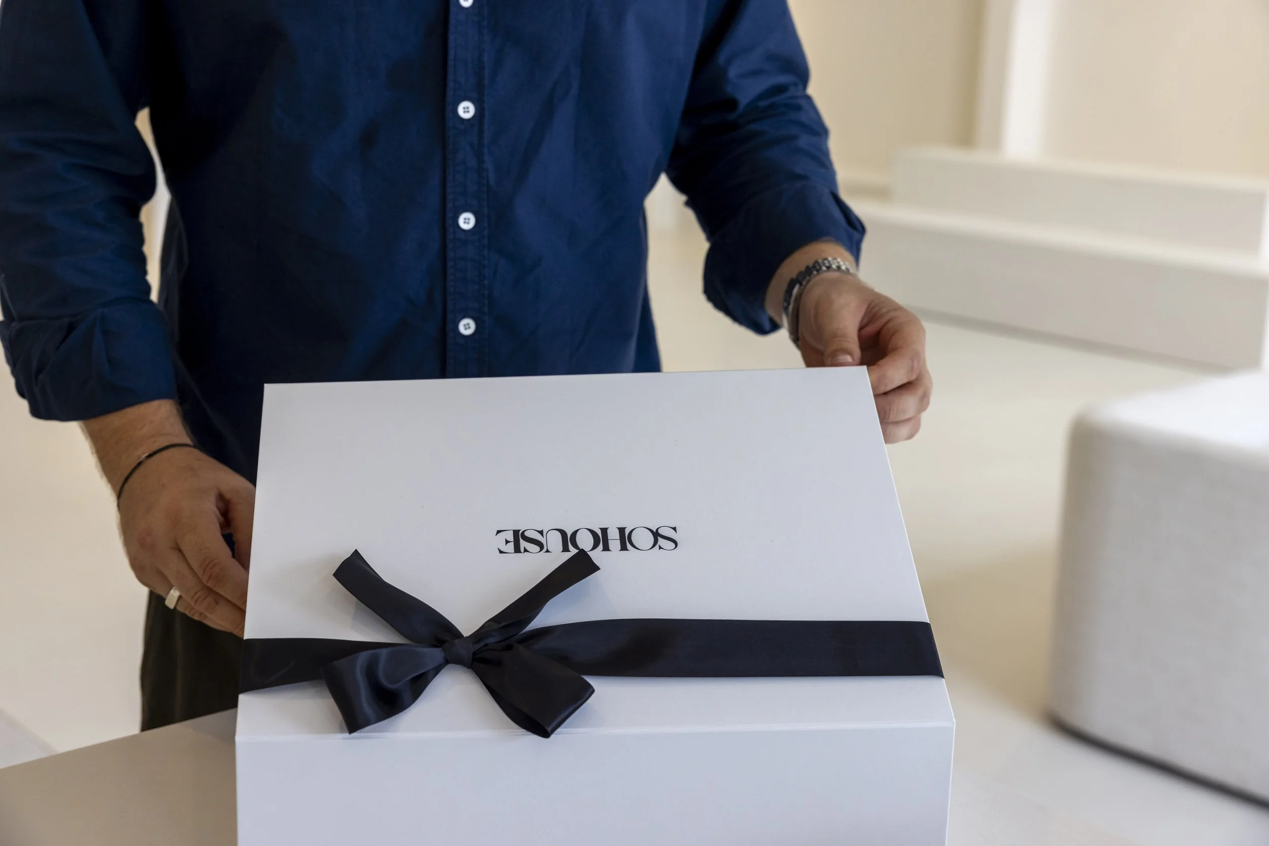 Brand Photography a Person in a blue shirt holding a white box with a black ribbon and text that reads 'SOHOUSE'.