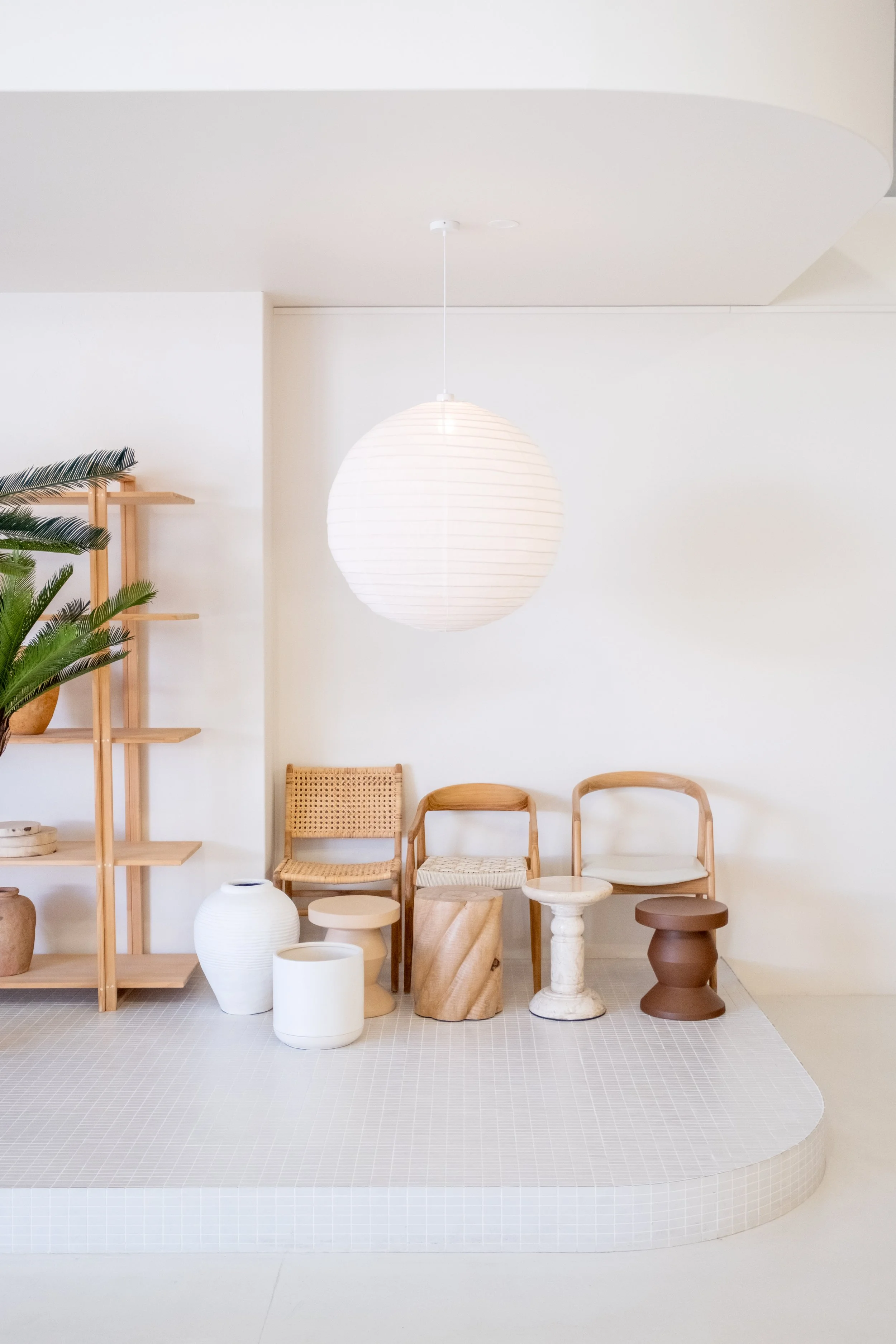 Minimalist interior with a white wall, hanging paper lantern, and a row of diverse wooden chairs and stools, alongside decorative pots and a wooden bookshelf with plants.