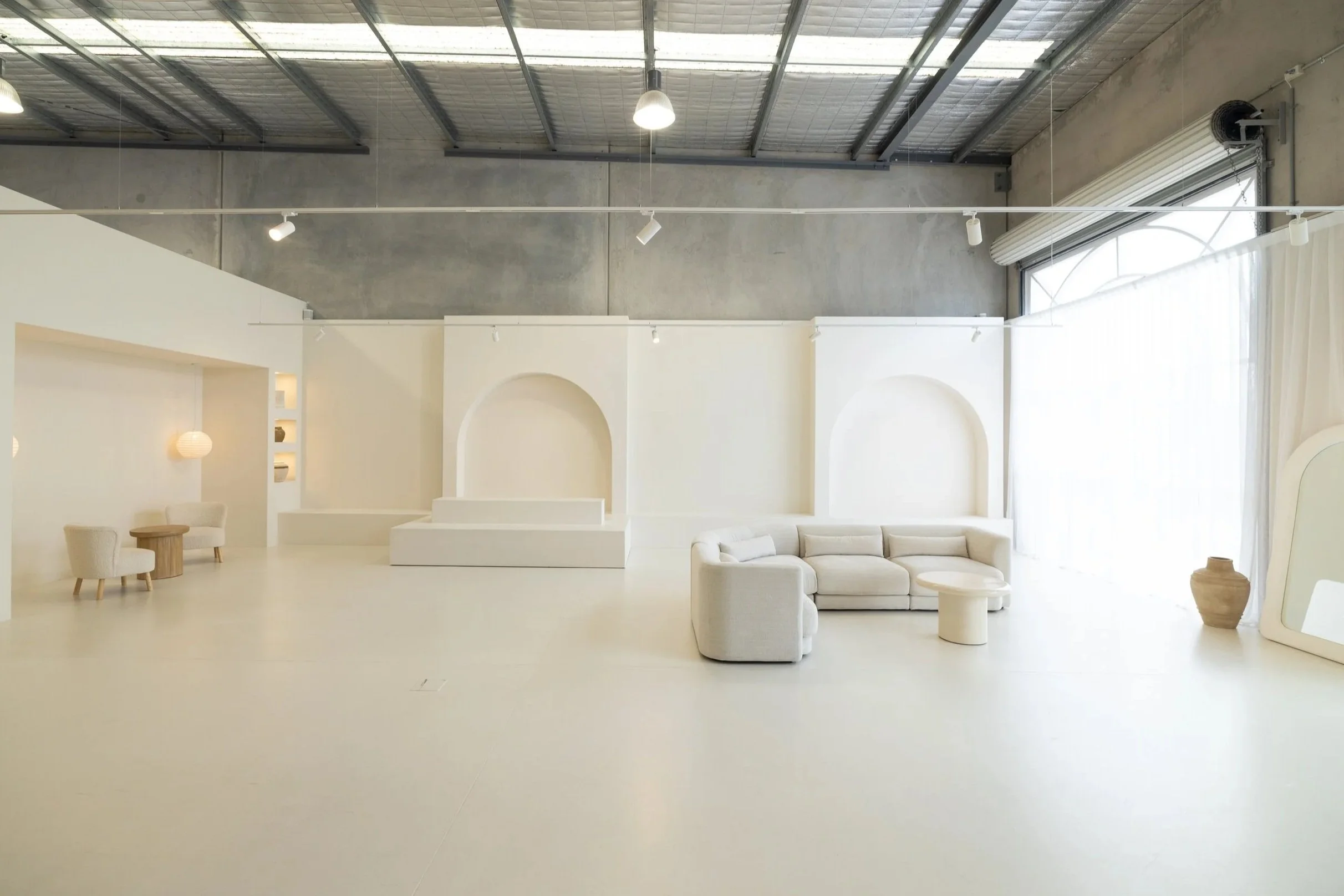 Luma Space Buderim is a spacious, modern interior with a high ceiling, concrete walls, and large windows. The room is decorated with white furniture, including a sectional sofa, chairs, and small tables, with minimalistic decor.