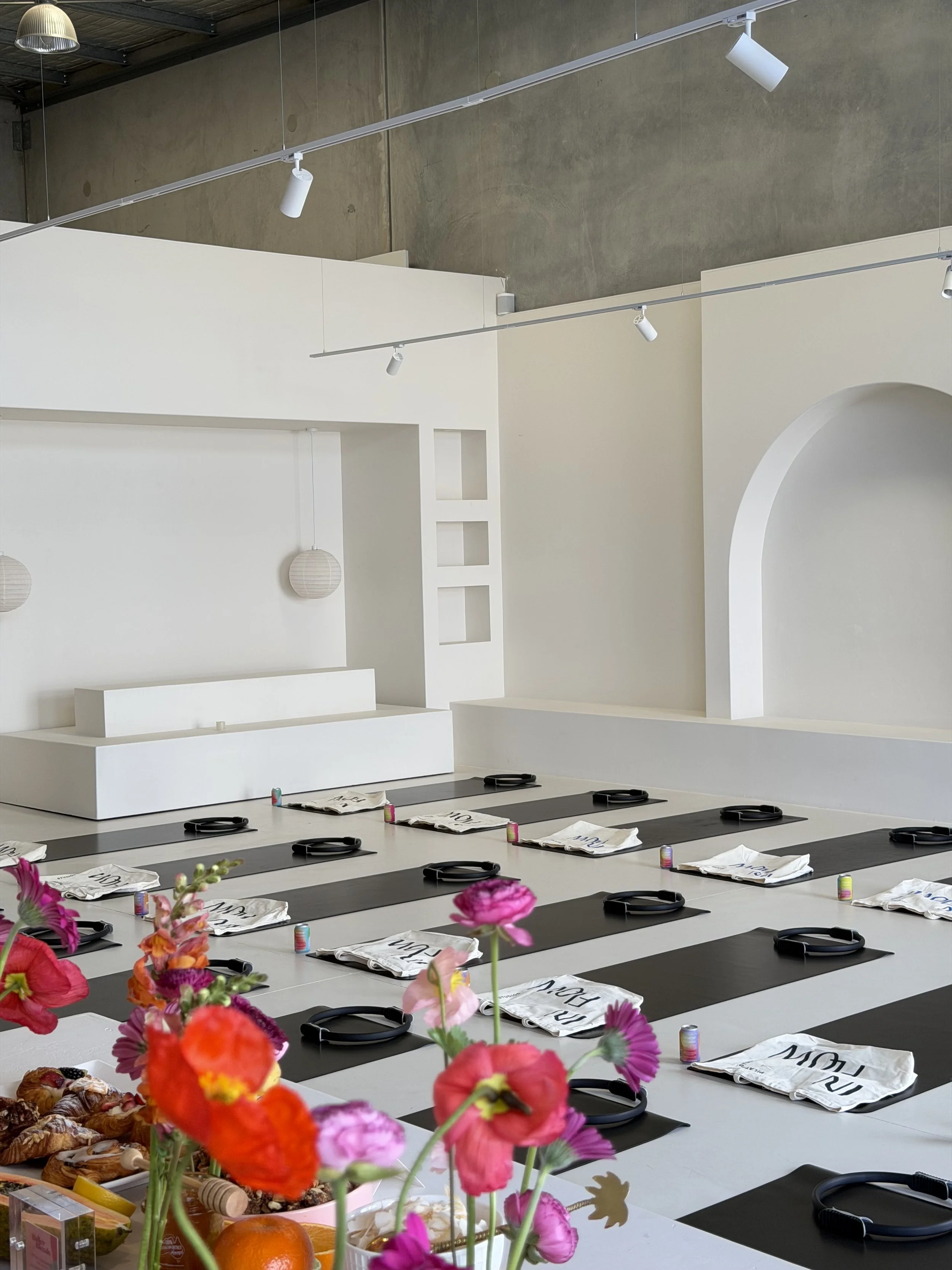 Luma space Buderim Sunshine Coast set up for a yoga or meditation class with black mats, rolled towels, and books or notepads on each mat, surrounded by colorful flowers, with a white wall, decorative squares, and a ceiling with track lighting.