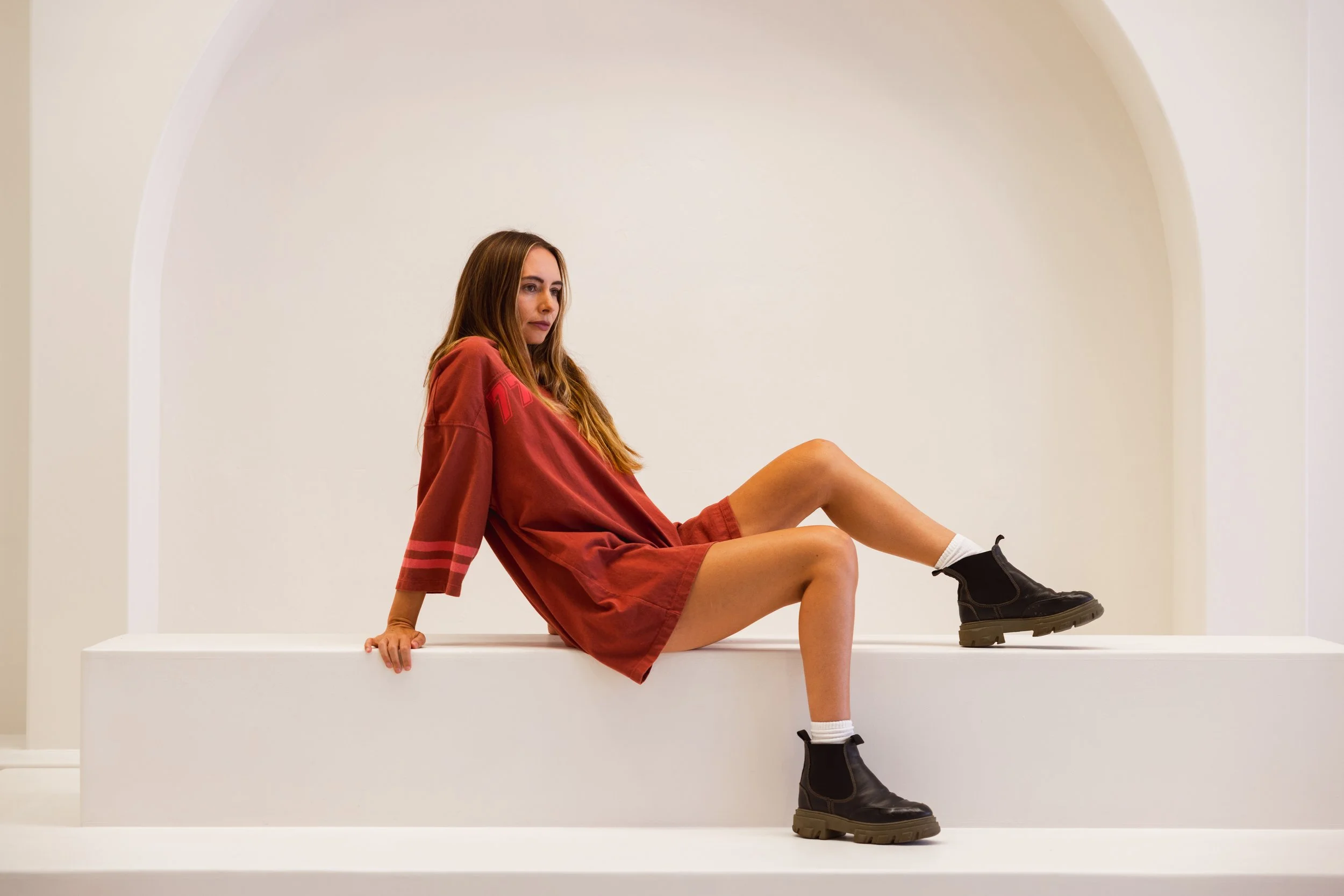 creative photography shoot in the Luma Space Sunshine Coast this is a woman with long brown hair sitting on a white platform inside a white cube, wearing a red hoodie, shorts, and black boots, with one leg bent and the other extended.