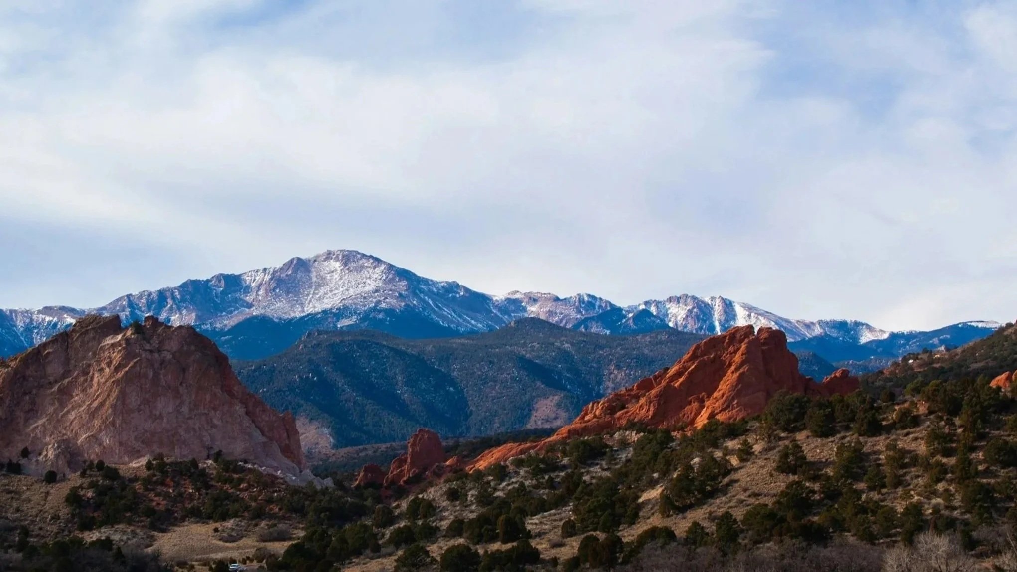 Mountain landscape with rocky red formations in the foreground, green trees, blue snow-capped mountains, and a light cloudy sky in the background.