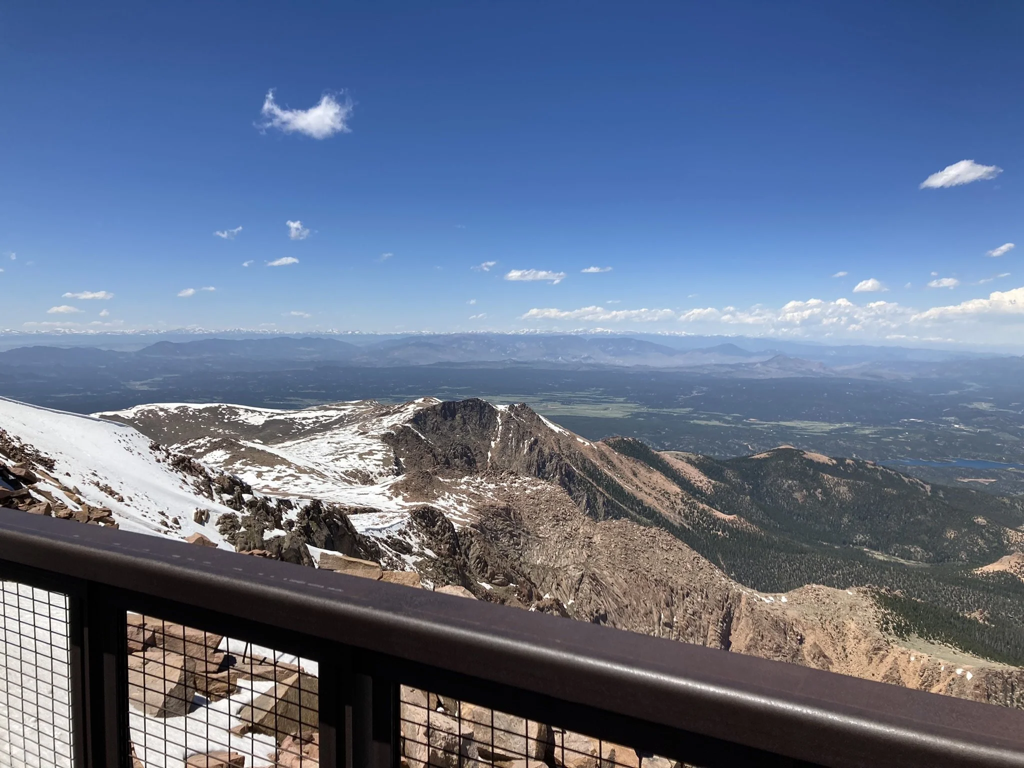 Mountain view from a lookout with snow-capped peaks, rocky ridges, a valley below, and a clear blue sky with a few scattered clouds.