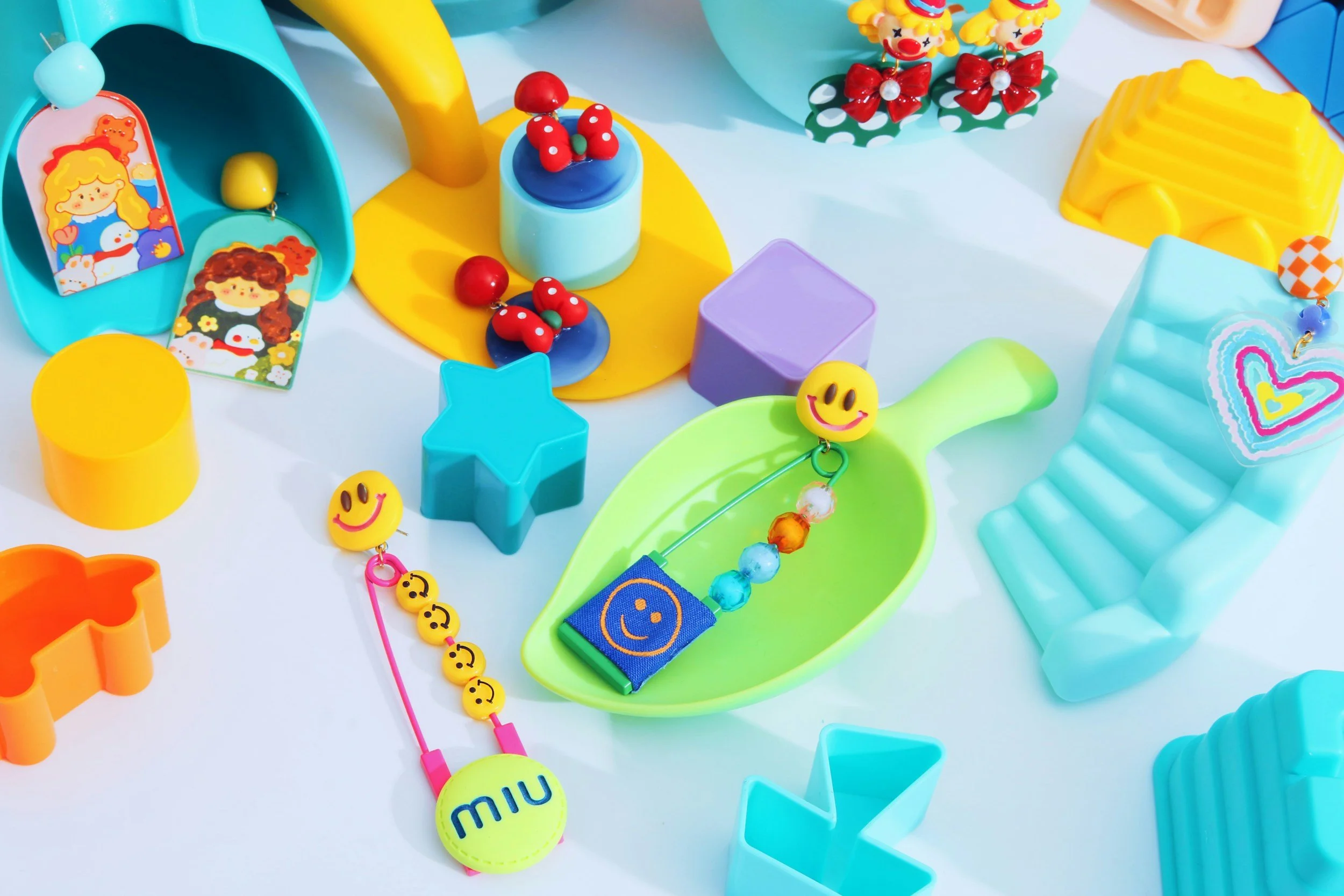 Colorful assortment of toys including smiley face pendants, animal-shaped buttons, geometric shapes, and decorative items arranged on a white surface.