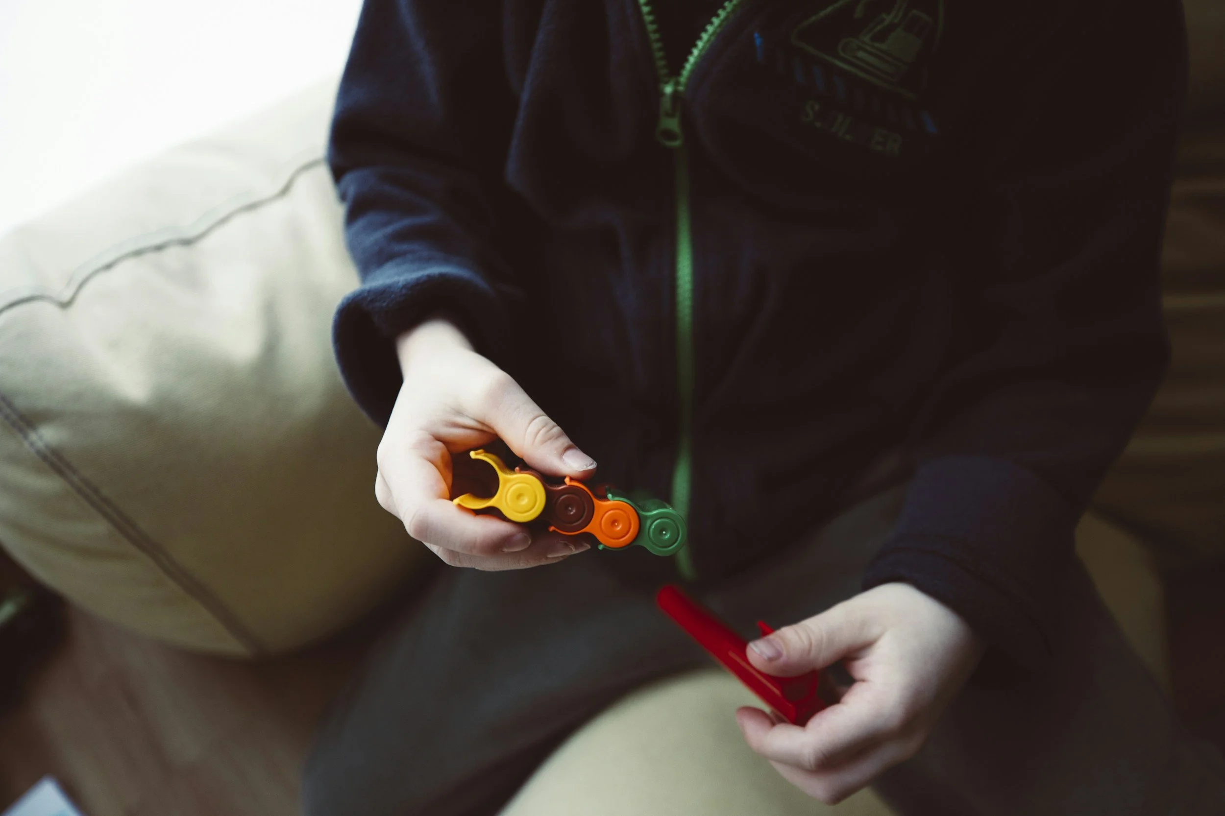 A person is holding a colorful fidget toy with yellow, orange, and green sections and a red pen in their other hand, sitting on a beige chair.