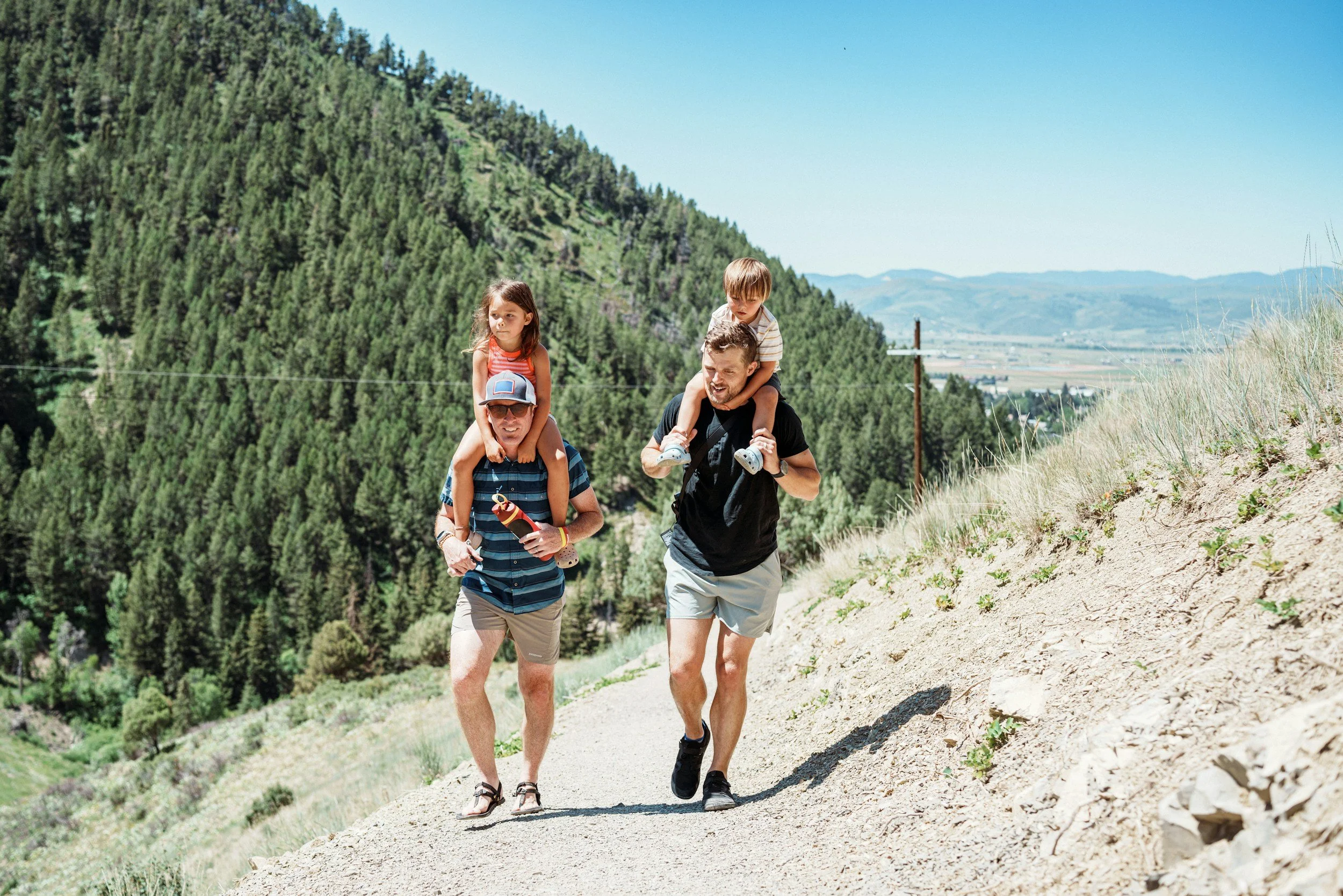 A family of four hiking on a dirt trail in a mountainous area with green trees and blue sky, with two children being carried on adult's shoulders.