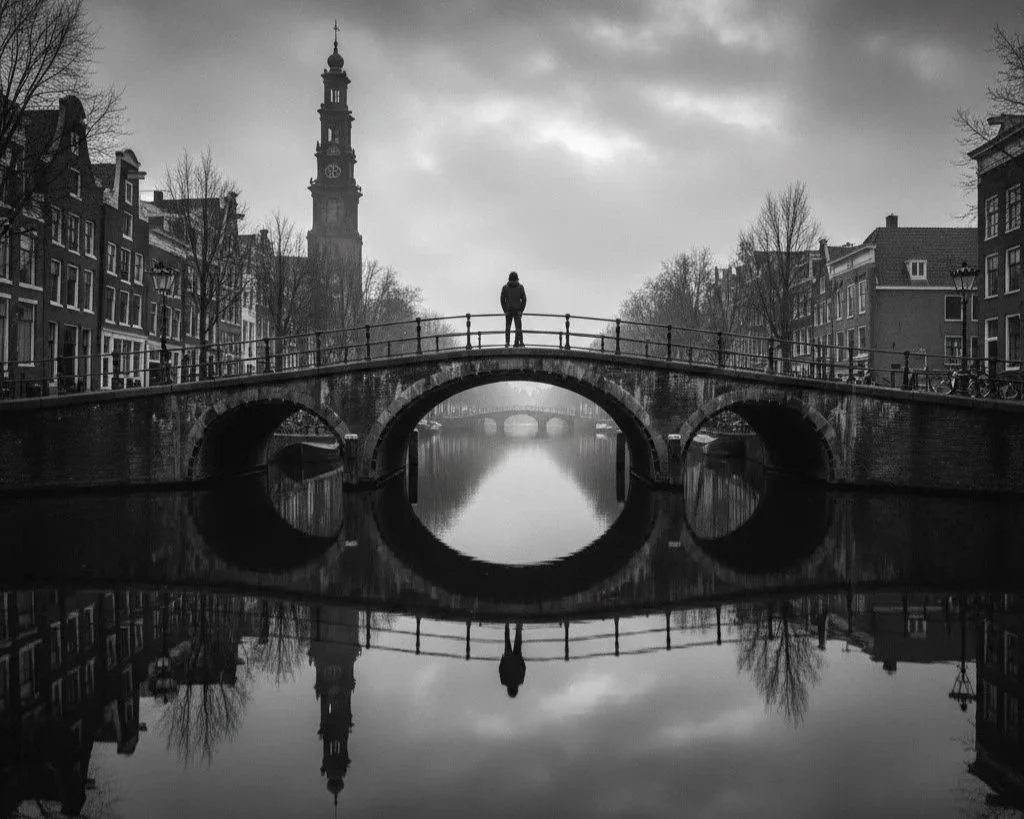 A black and white photo of a city canal with a bridge and a person standing on it, with historic buildings and a clock tower in the background.