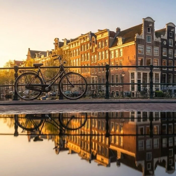 A bicycle parked along a canal railing in a European city with historic brick buildings, reflecting in the water at sunset.