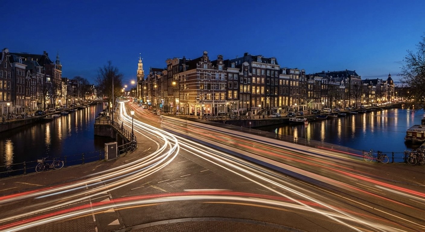 Night view of a city canal with illuminated buildings, light trails from moving vehicles on the street, and reflections of lights on the water.