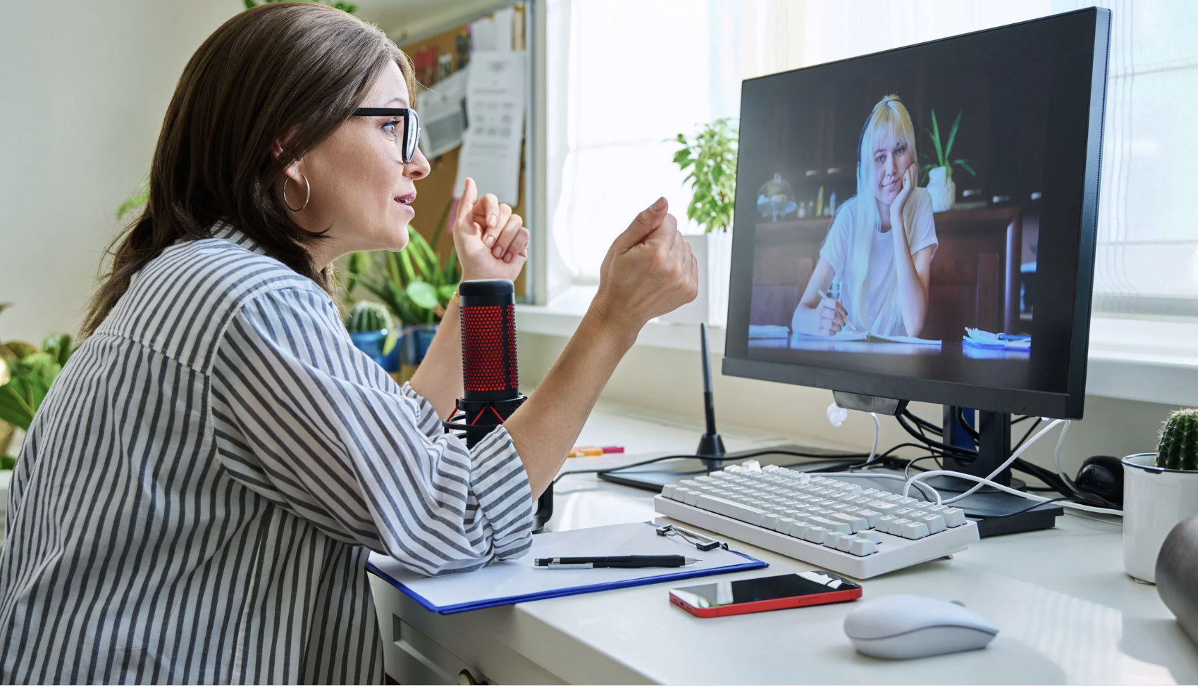 Woman with glasses and striped shirt participating in a video call with girl on computer screen, surrounded by plants, a microphone, and various office supplies.