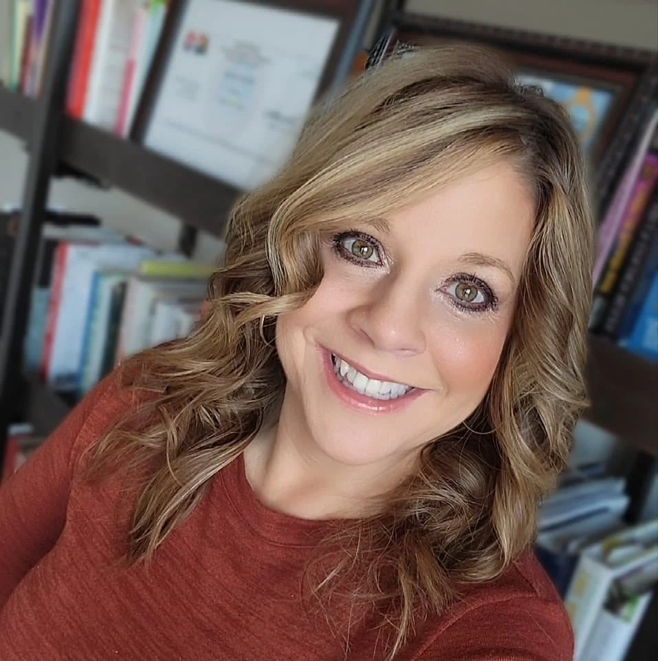 Smiling woman with wavy light brown hair and hazel eyes, wearing a rust-colored top, in front of a blurred bookshelf.