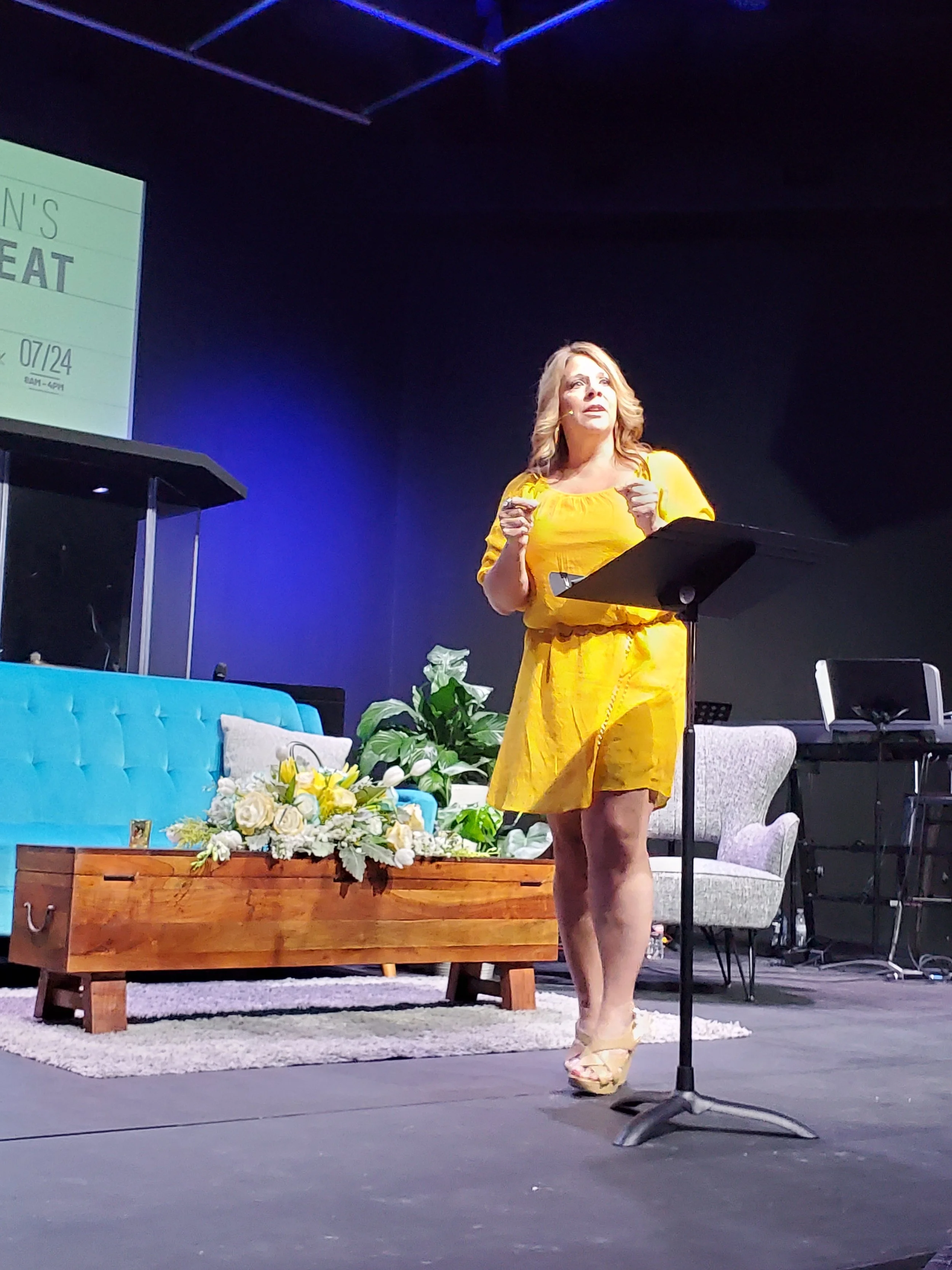 A woman in a yellow dress standing on stage delivering a speech, with a lectern and floral arrangement behind her.