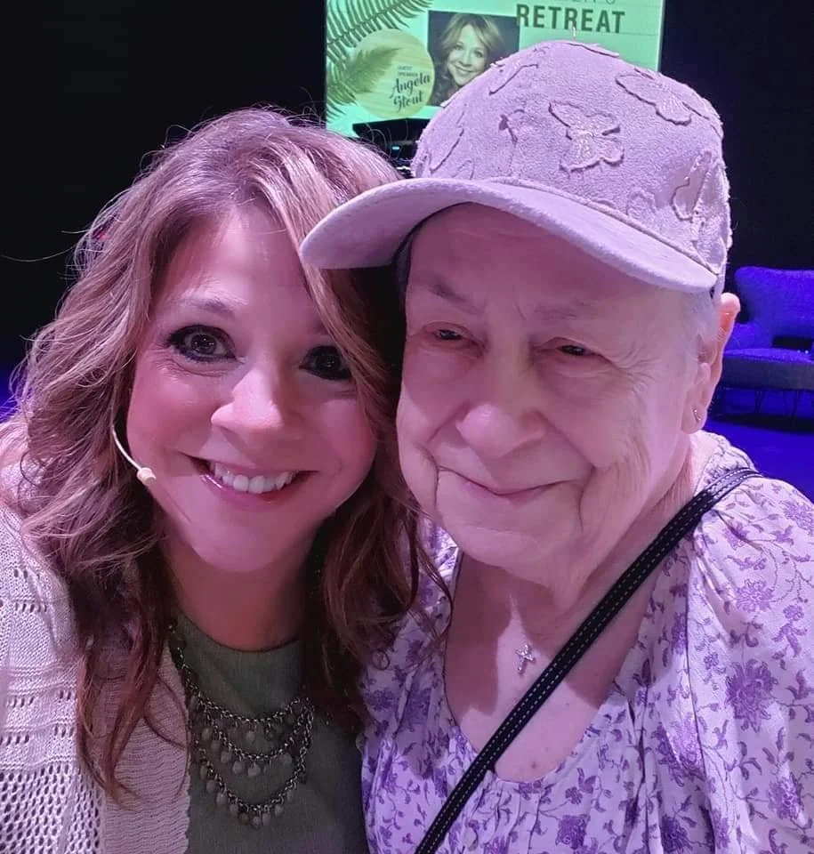 A smiling woman with light reddish wavy hair taking a selfie with an elderly woman wearing a light-colored cap with floral designs, in a dimly lit indoor setting with a background screen showing a woman's photo and text.