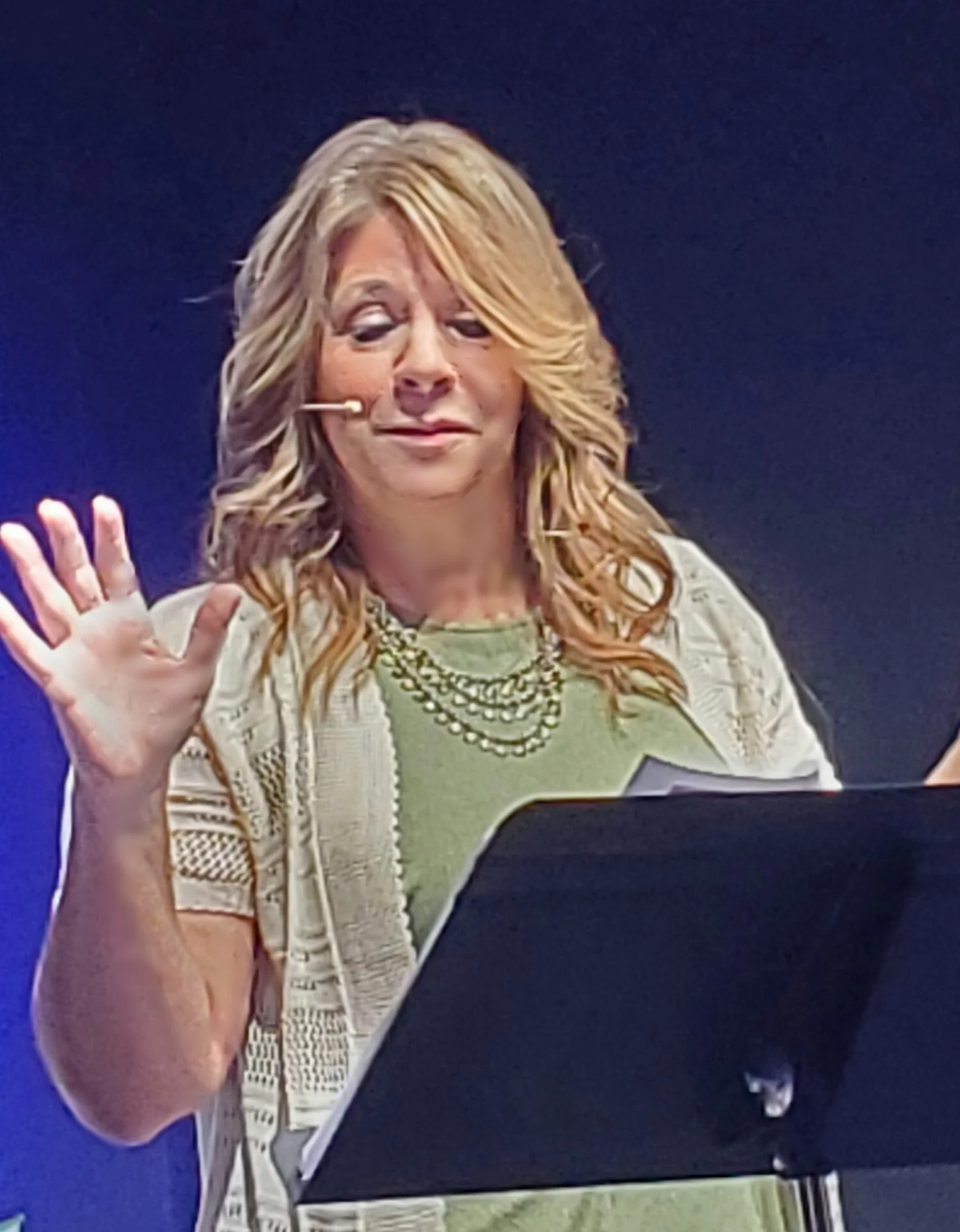 A woman with blond hair wearing a green top, a beige cardigan, and a layered pearl necklace, speaking on stage with a microphone headset, waving her hand.