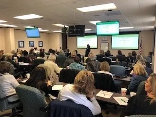 Group of people attending a conference or seminar in a large room with multiple screens and a presenter at the front.
