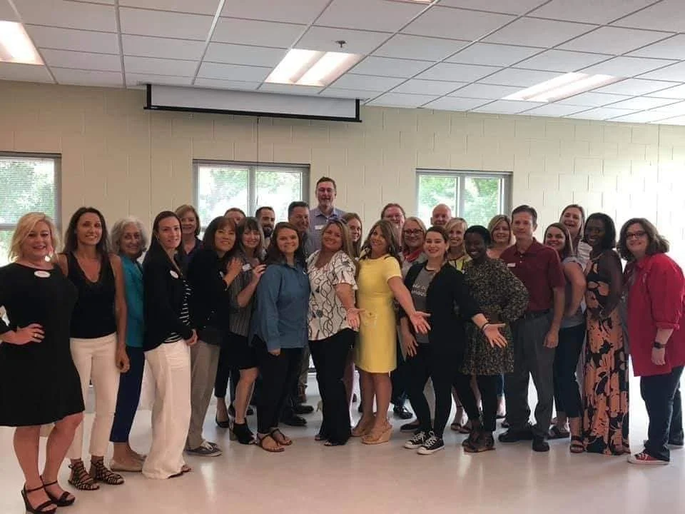 Group of diverse women and men posing together in a classroom or community center, with some smiling and gesturing.