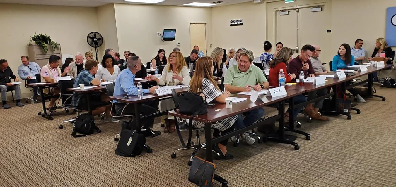 A large room filled with people seated at long conference tables during a meeting or conference, engaging in discussions, with some taking notes and others listening.