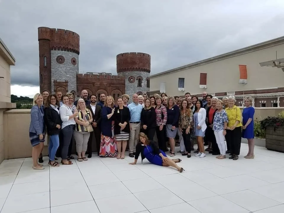 A group of people posing on a rooftop terrace with a castle-style building in the background. The group includes men and women, some holding drinks, with one woman lying on the ground in front.