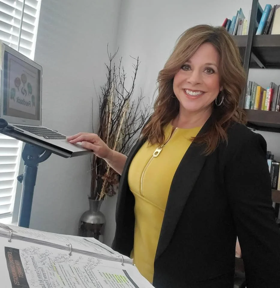A woman with shoulder-length light brown hair, wearing a yellow top and black blazer, smiling while holding a laptop on a stand. She is in a room with a bookshelf, a tall vase with dried branches, and a window with shade blinds.