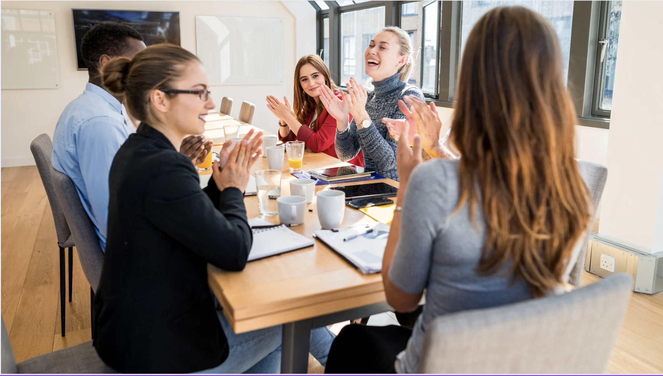 A group of people sitting around a conference table, clapping and smiling during a meeting in a bright room with large windows.