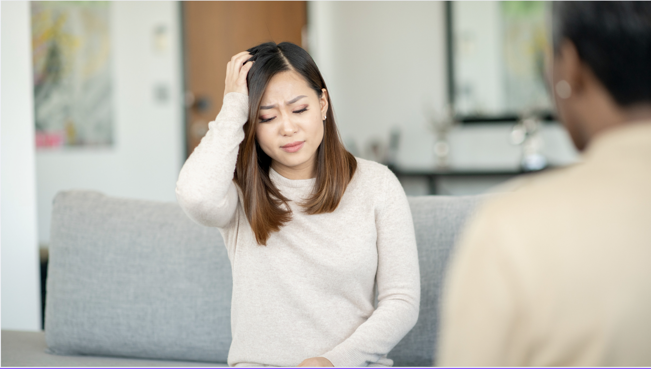 A young woman with brown hair in a beige sweater sitting on a gray couch with her hand on her head, appearing distressed or confused during a counseling session.